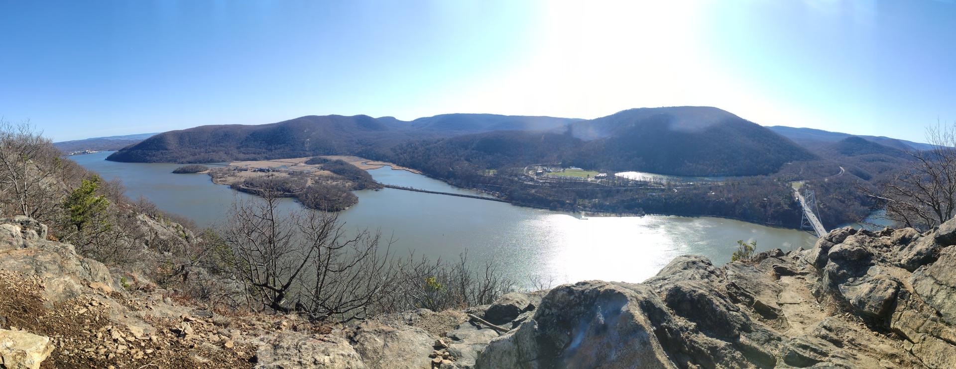 Sunny panoramic view of a wide river, distant bridges, and rocky foreground.