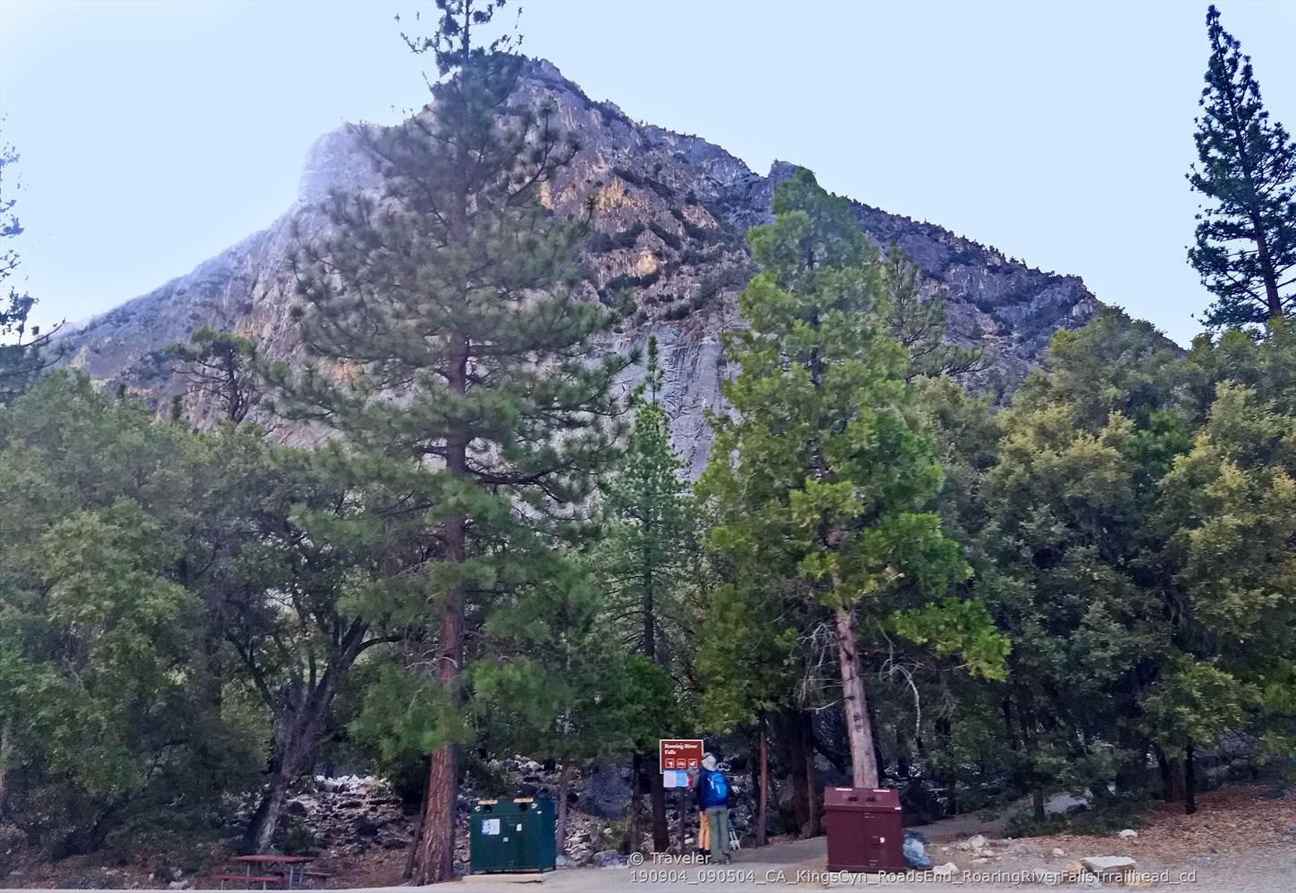 A person stands near bins and a sign below a massive rocky peak.