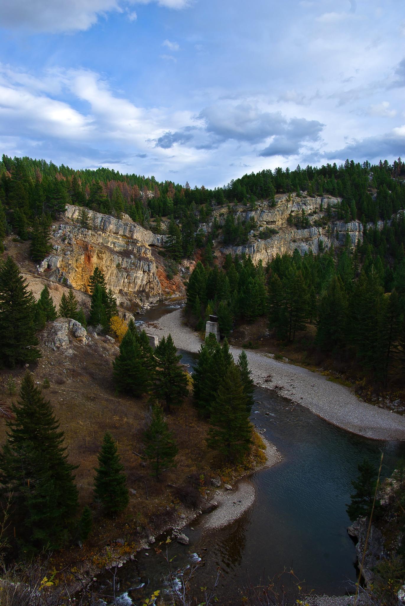 River flowing through a forested canyon with colorful rocky cliffs under cloudy sky.