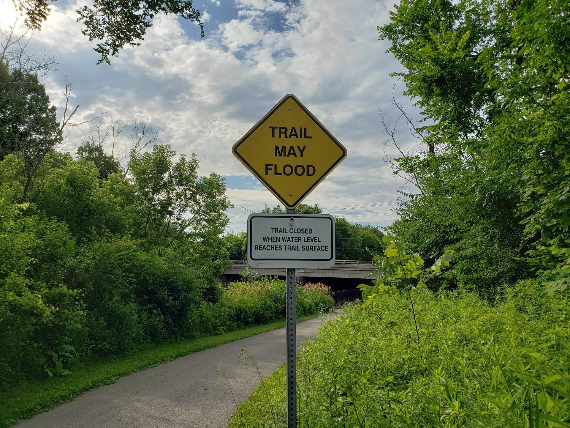 Flood warning sign on a paved trail surrounded by green trees.