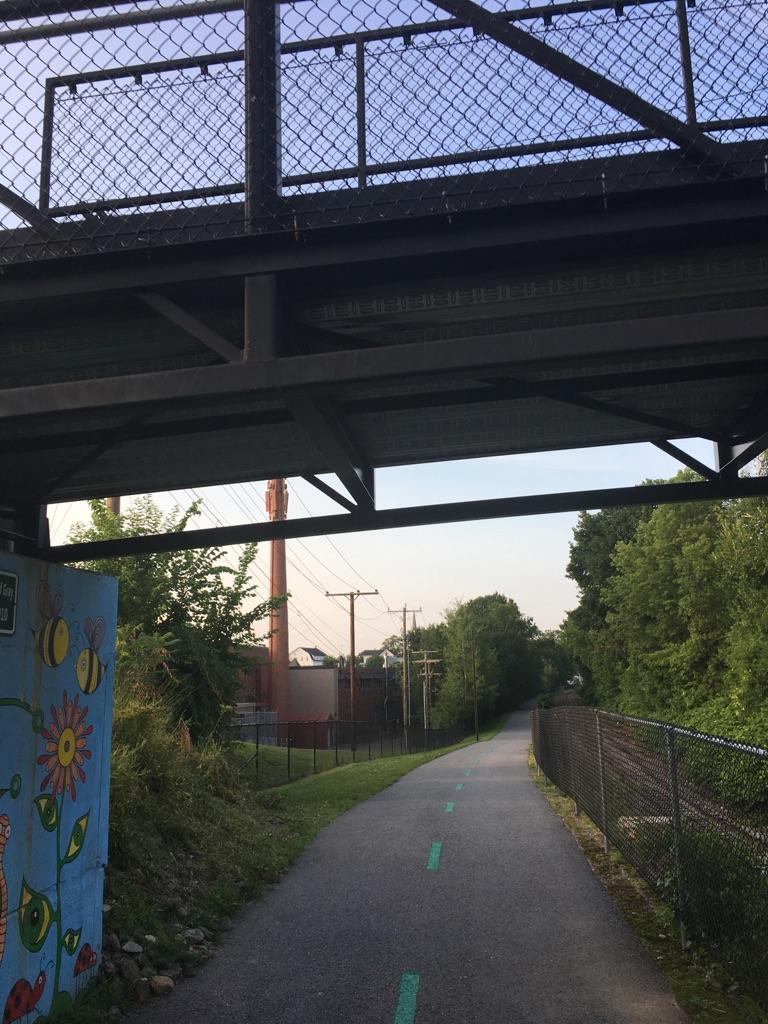 Paved path under a metal bridge, leading into a tree-lined area.