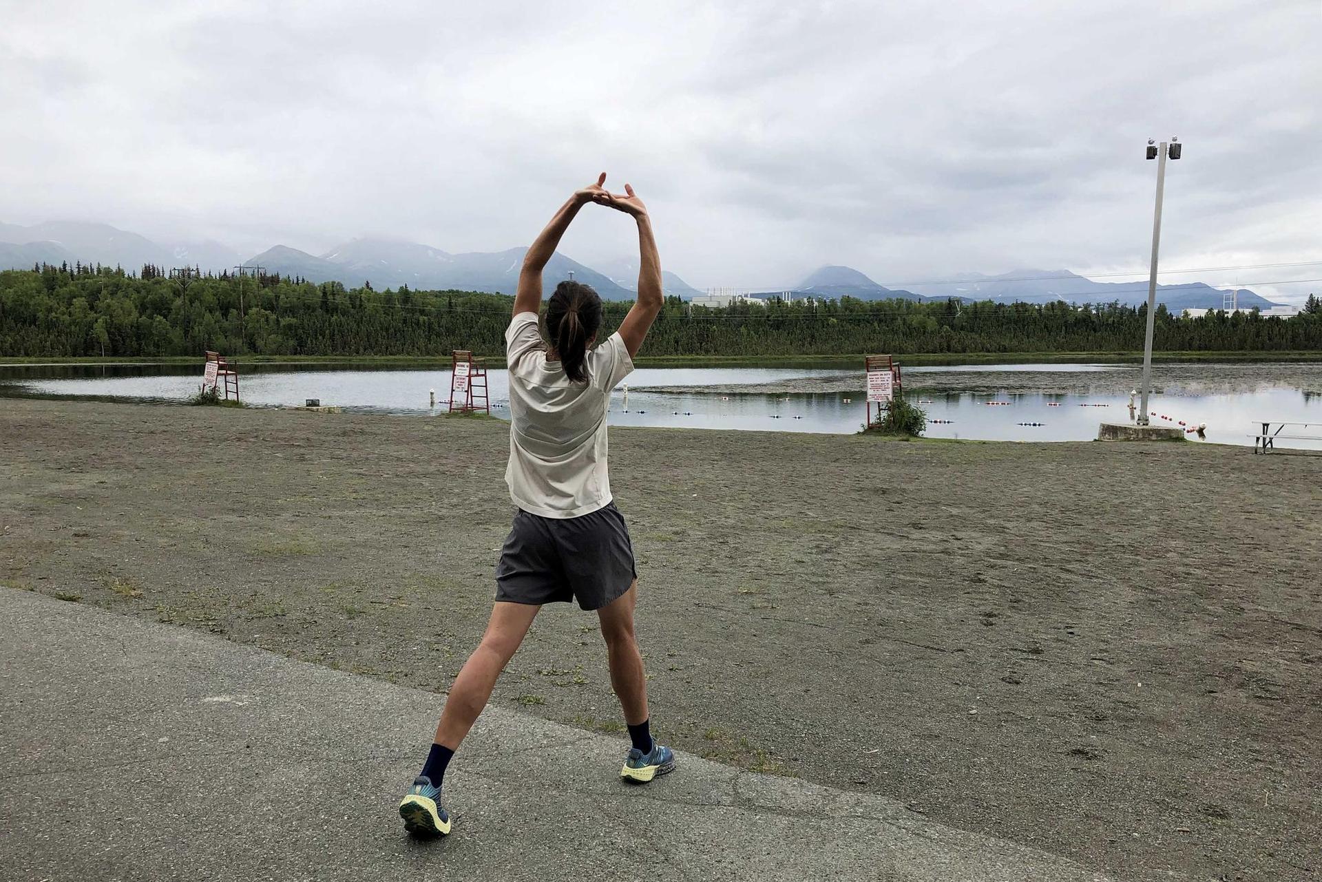 Person stretching by a lake with mountains under an overcast sky.