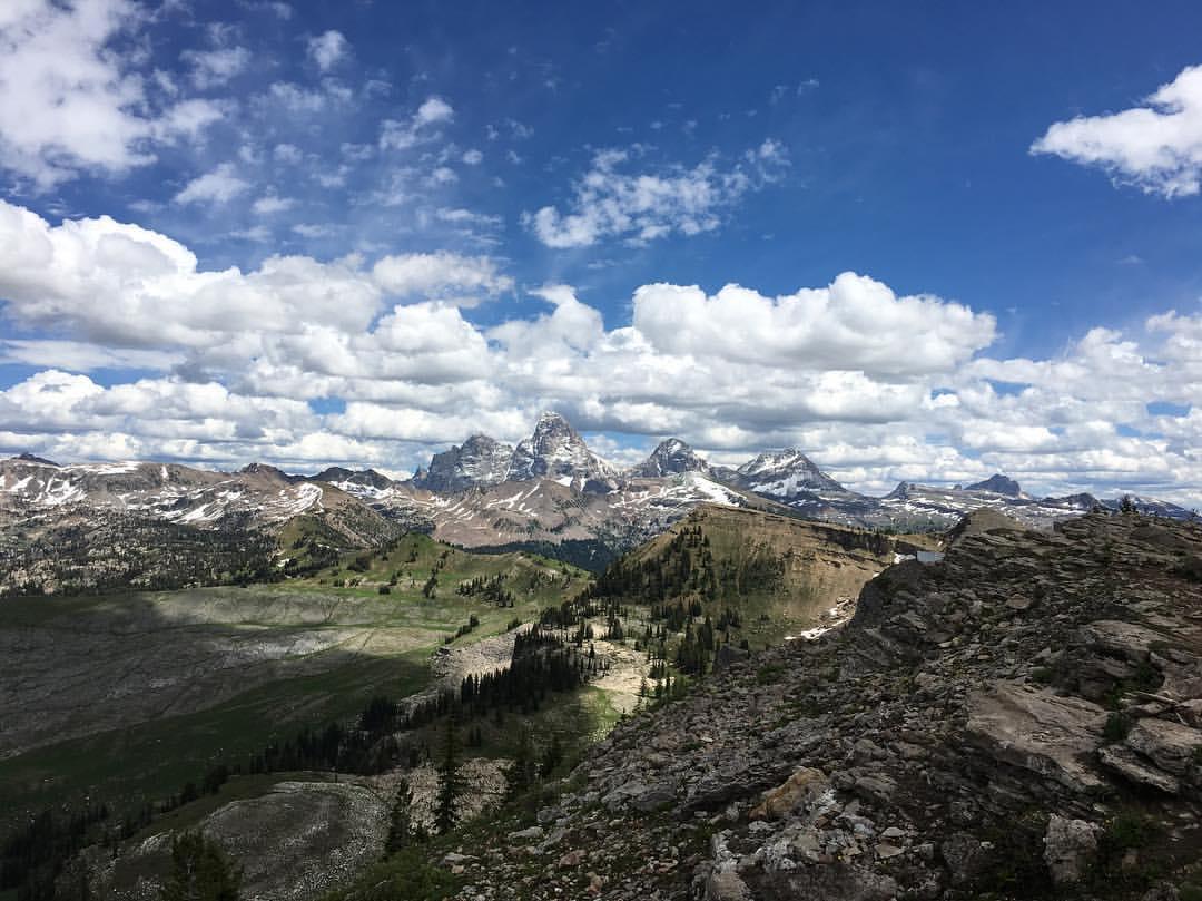 Panoramic view of majestic snow-capped mountains, green valleys, and a rocky foreground under a cloudy sky.
