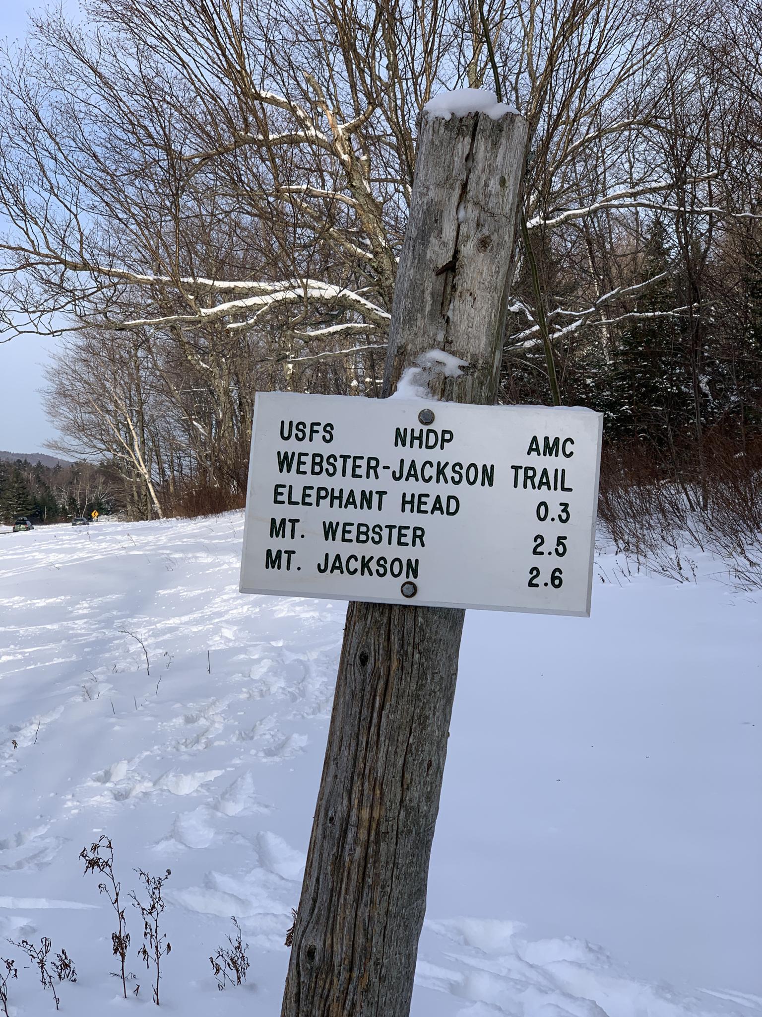 A wooden trail sign stands in deep snow on a bright winter day.