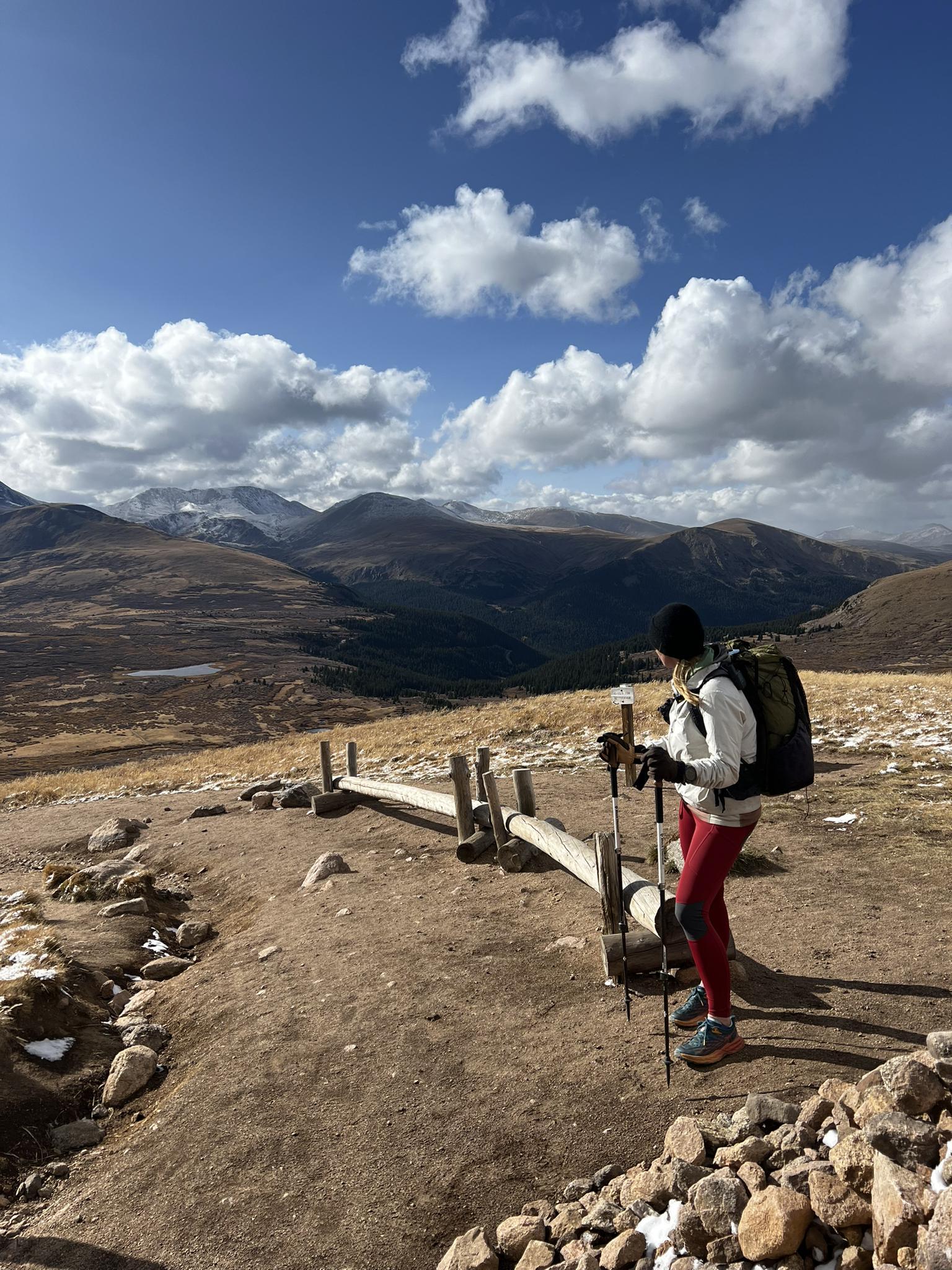 Hiker with poles overlooks a vast mountain landscape under a bright, cloudy sky.