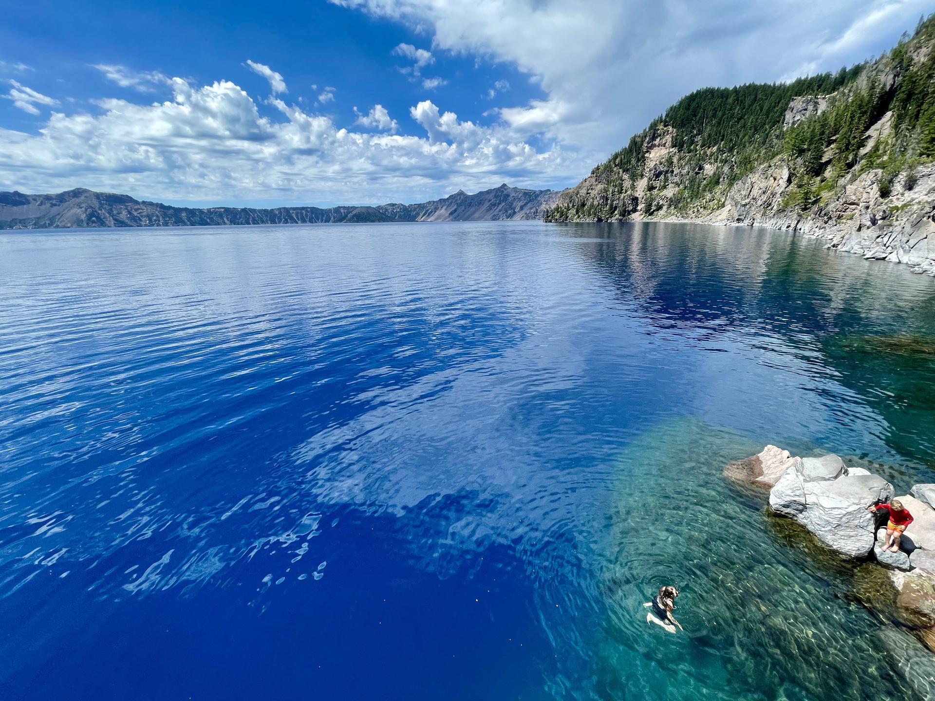 Clear blue lake with a swimmer near rocks, steep forested cliffs in background.