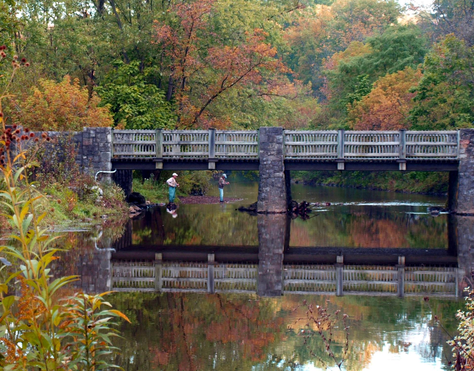 Two people fishing under a wooden bridge with autumn trees reflecting in the river.