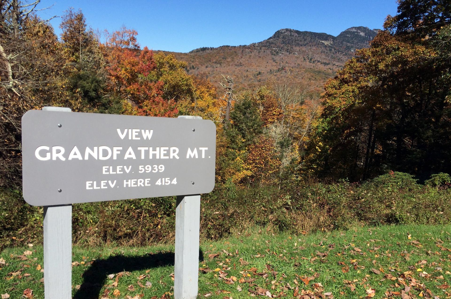 A gray sign in front of a mountain landscape with vibrant autumn foliage.