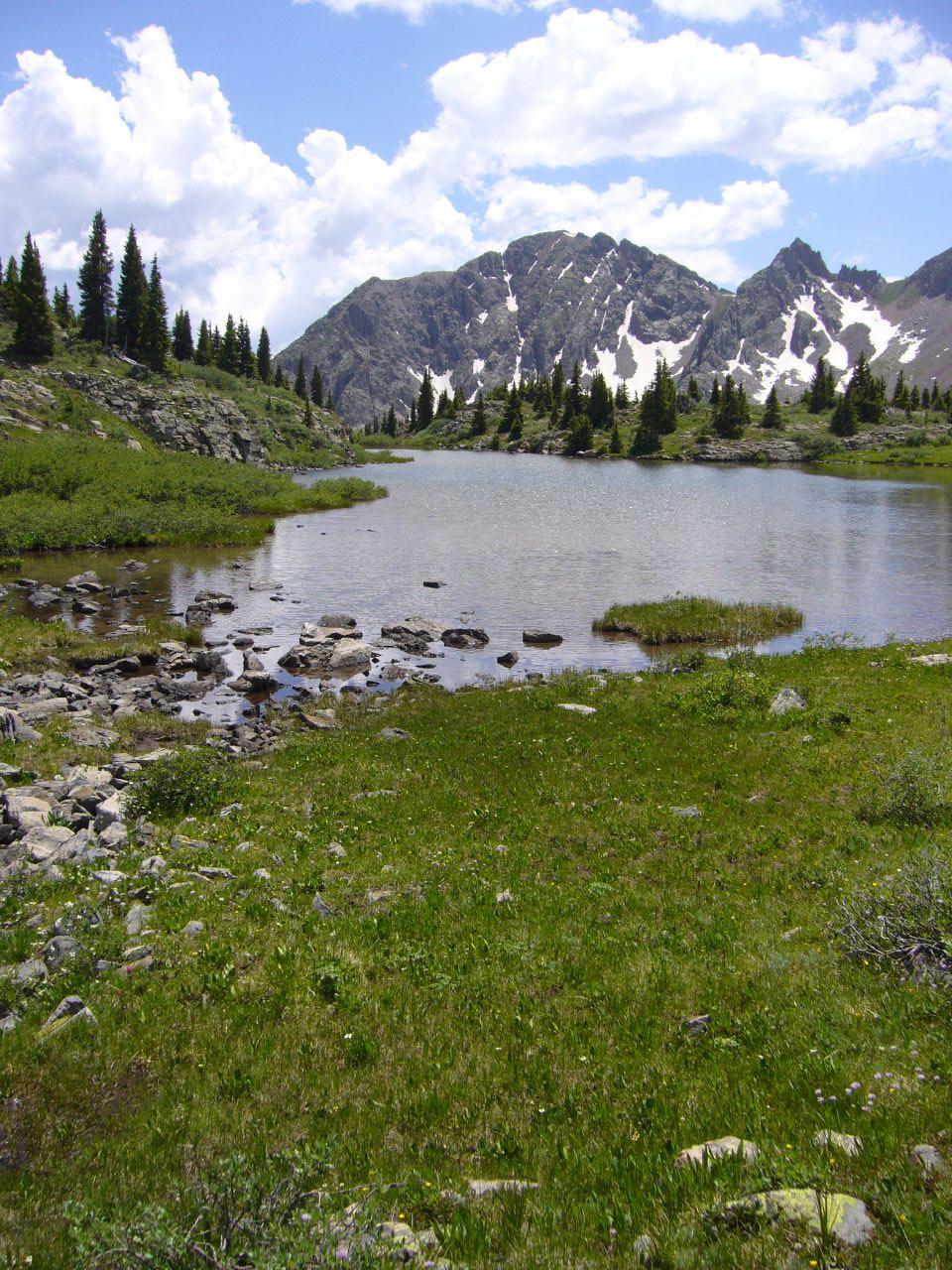 Serene mountain lake with rocky shore, green grass, and snow-capped peaks.