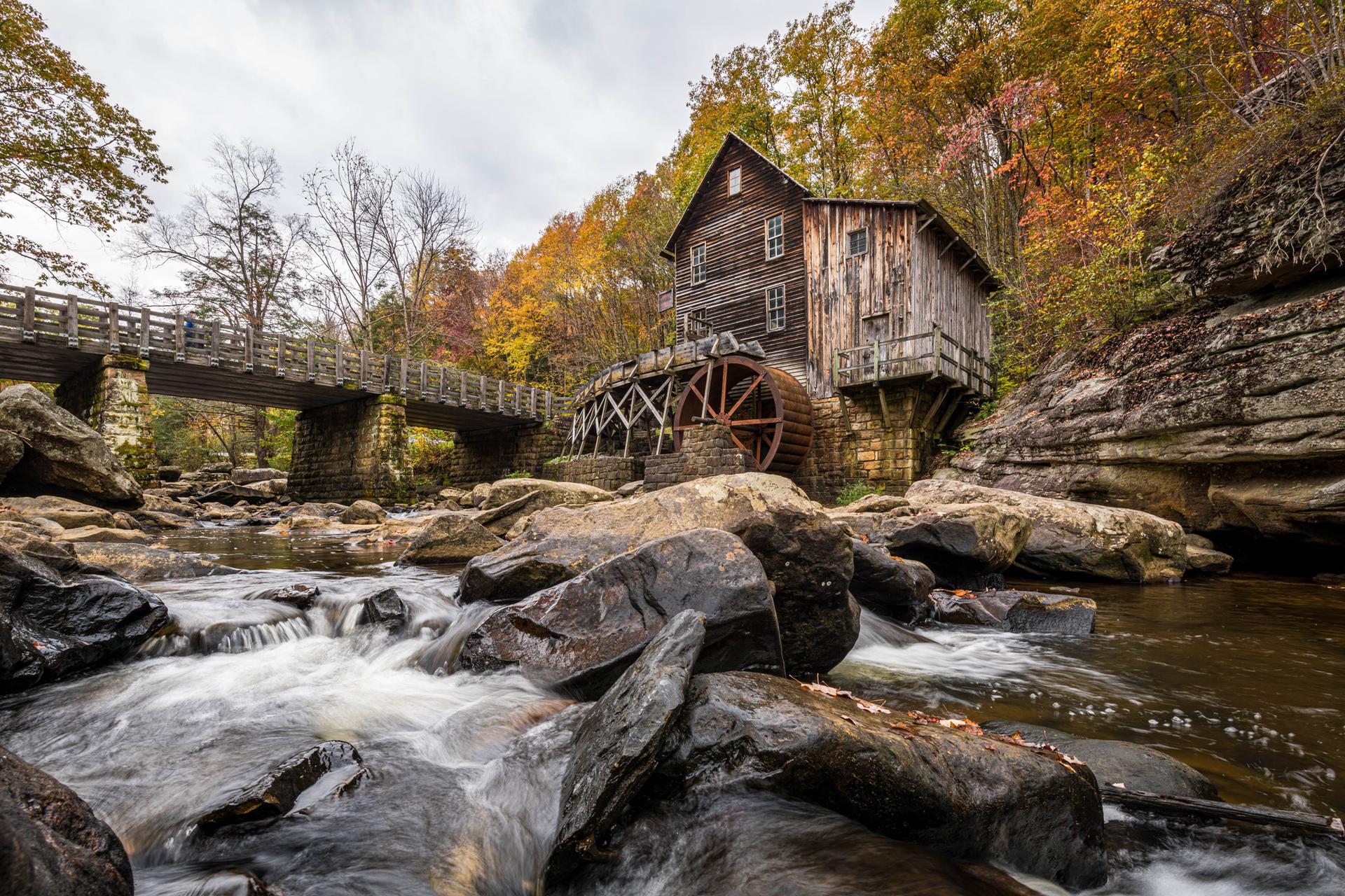 Historic wooden mill with water wheel by a rocky river and autumn trees.