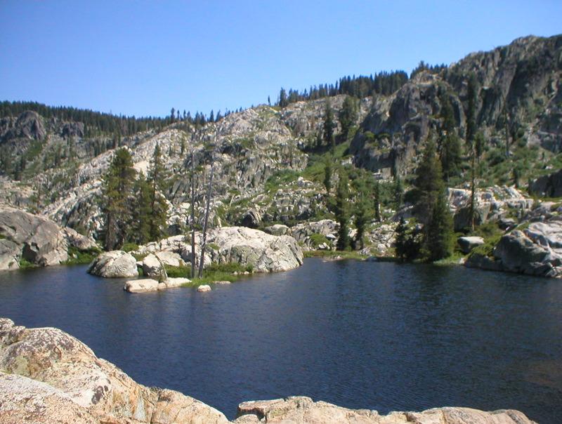 Calm lake water surrounded by large rocks and pine trees on a sunny day.