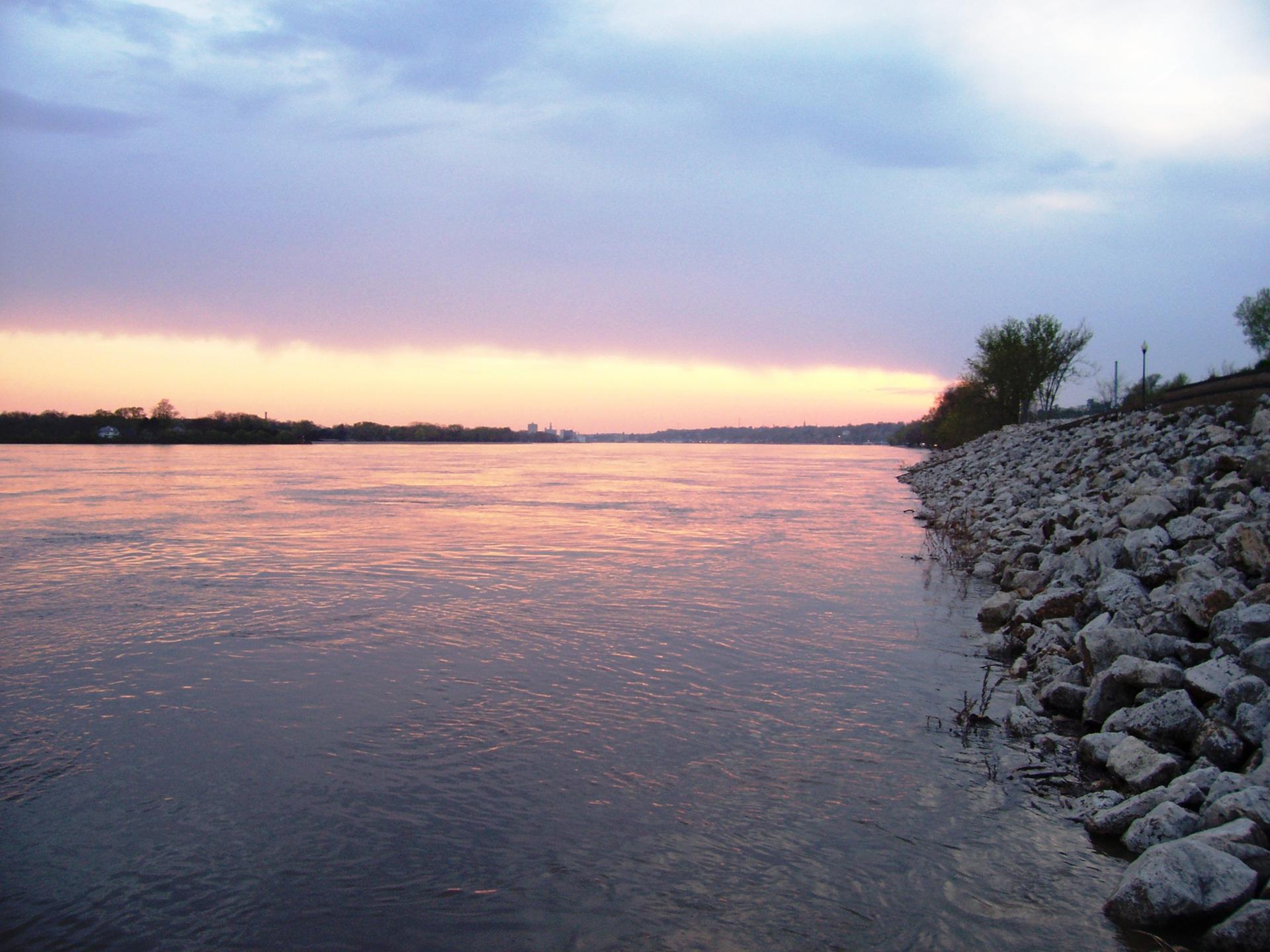 Wide river reflecting a colorful sunset sky with a rocky bank.