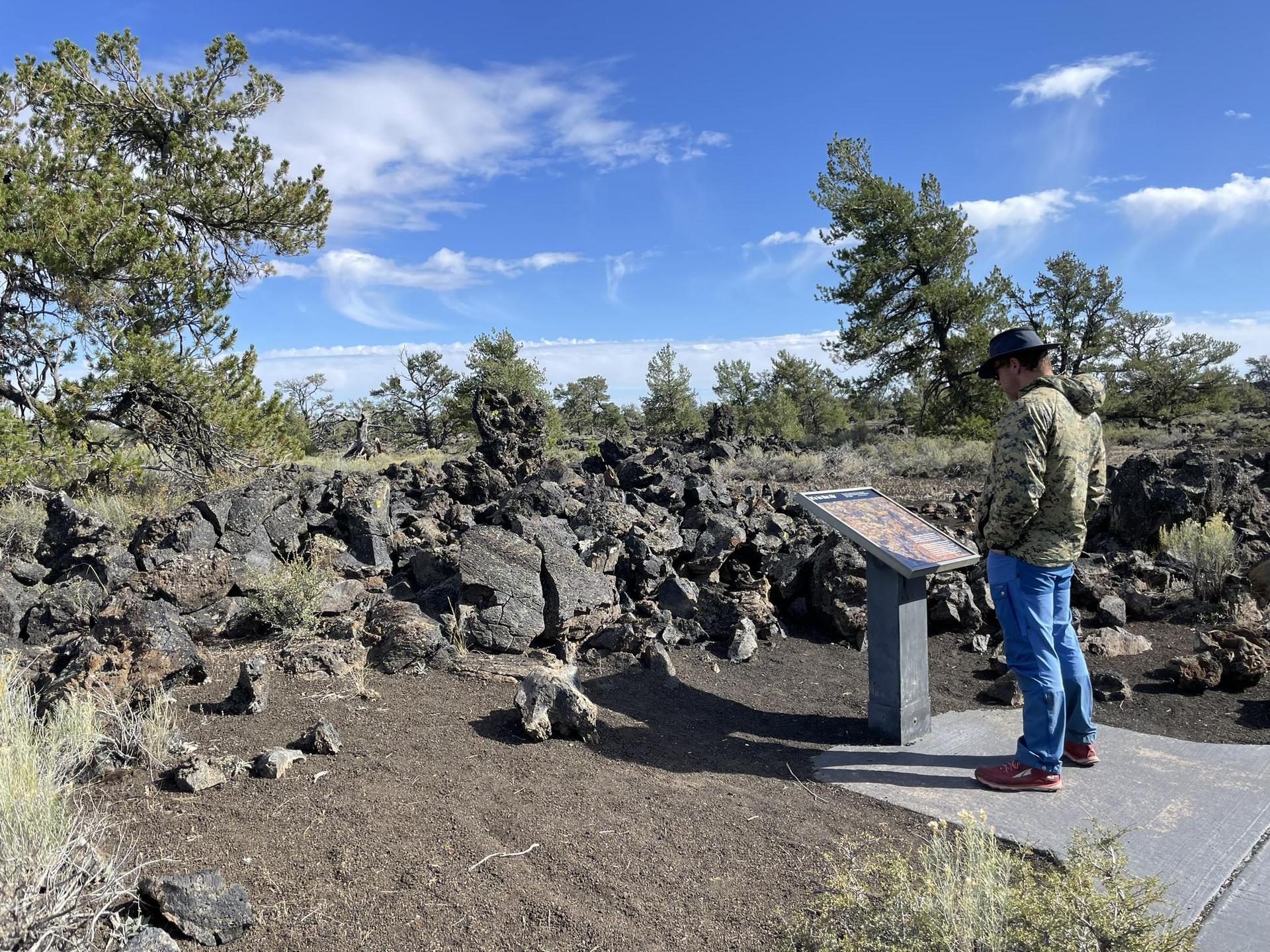 Man in hat reads an outdoor sign in a rocky, tree-filled landscape.