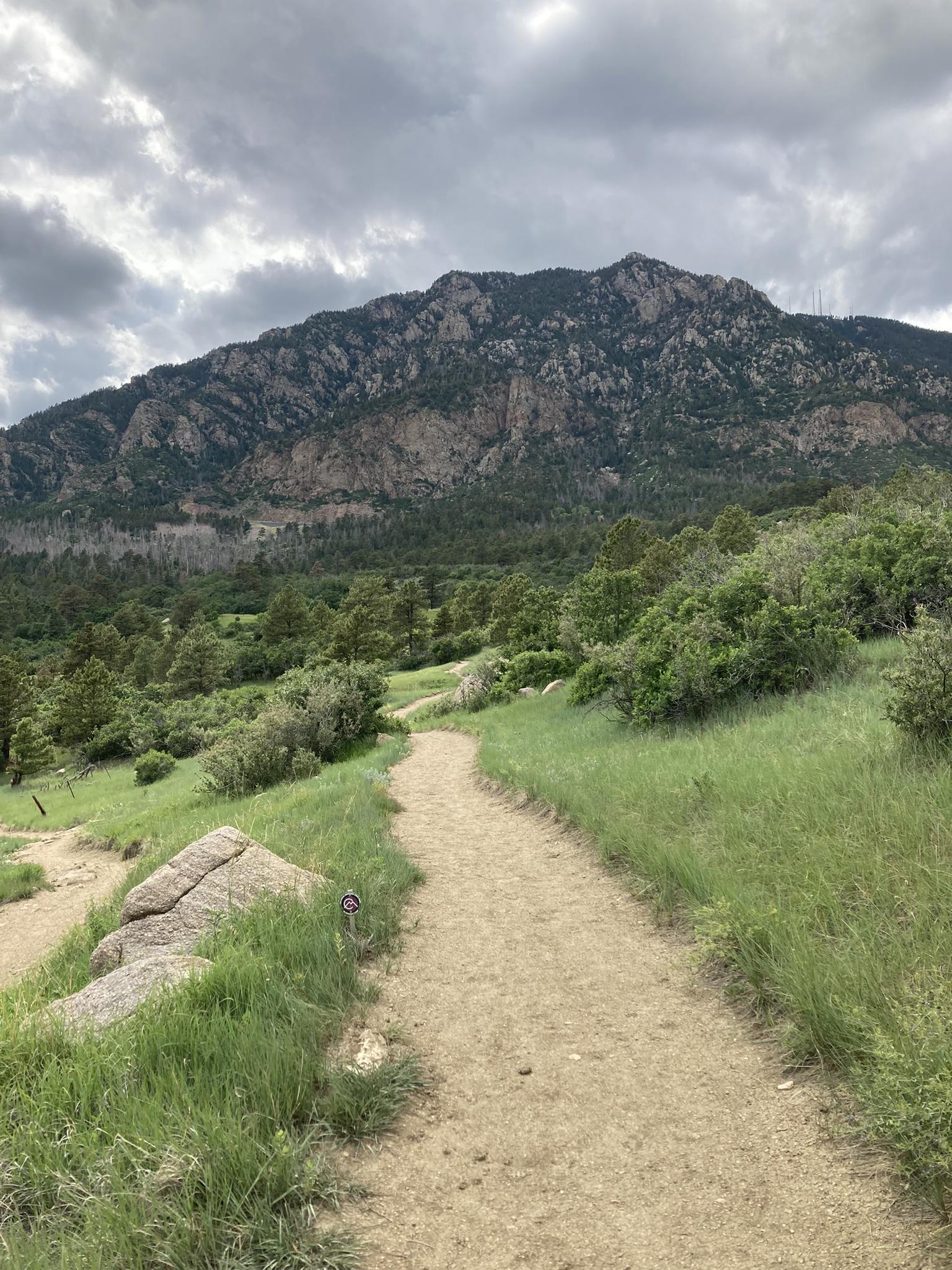 A dirt path winds through green grass towards rocky mountains under a cloudy sky.