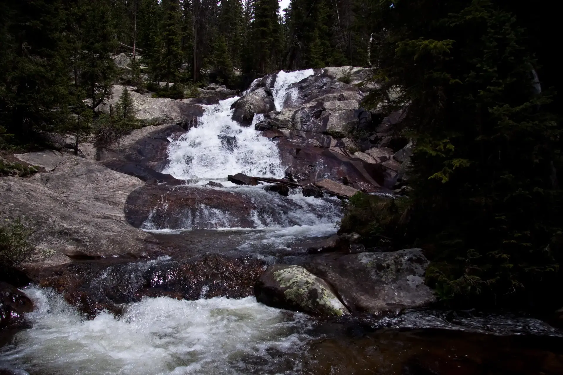 A multi-tiered waterfall cascades over dark rocks, surrounded by dense green trees.