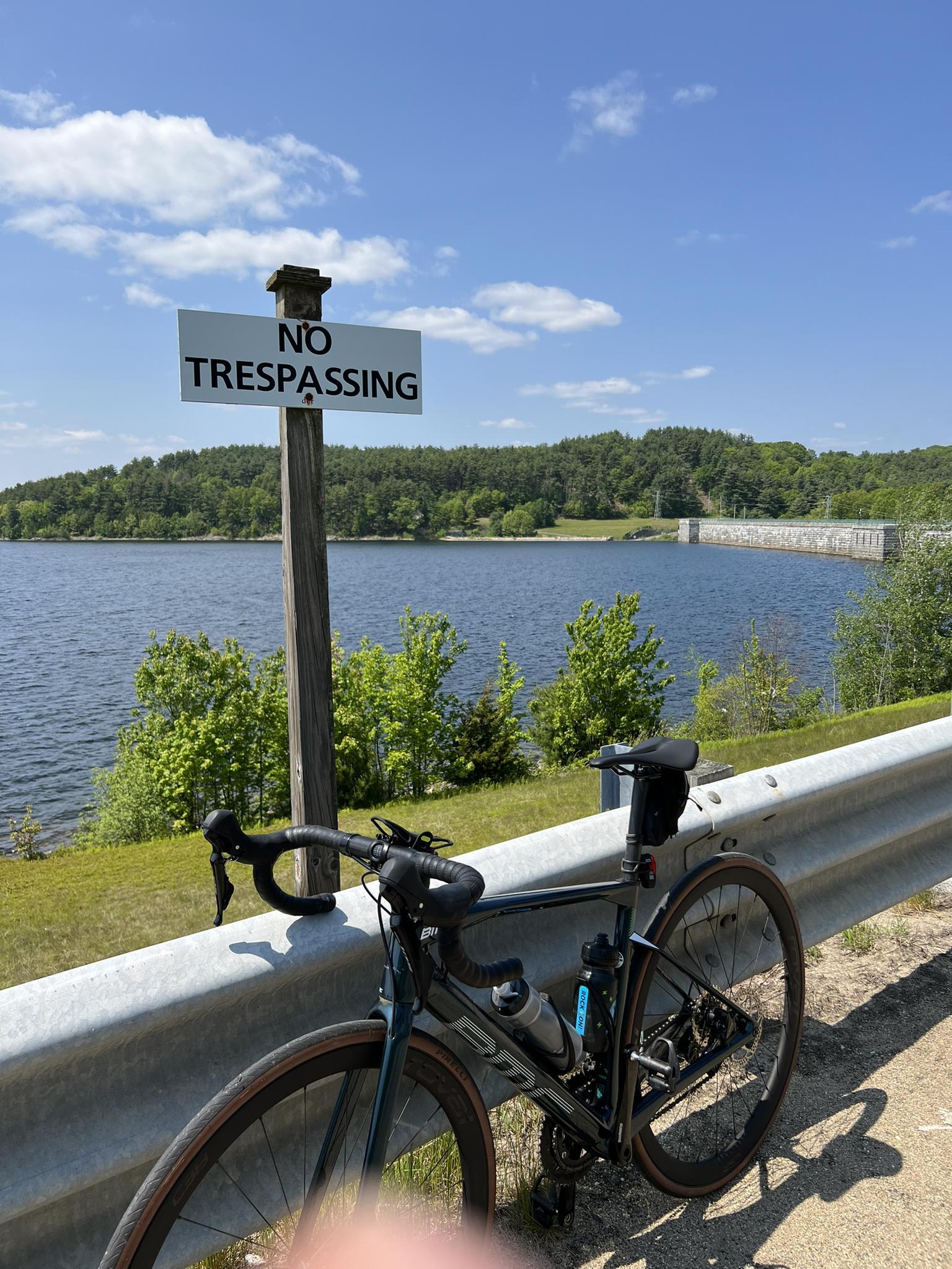 Bicycle parked by a 'No Trespassing' sign overlooking a large lake and dam.