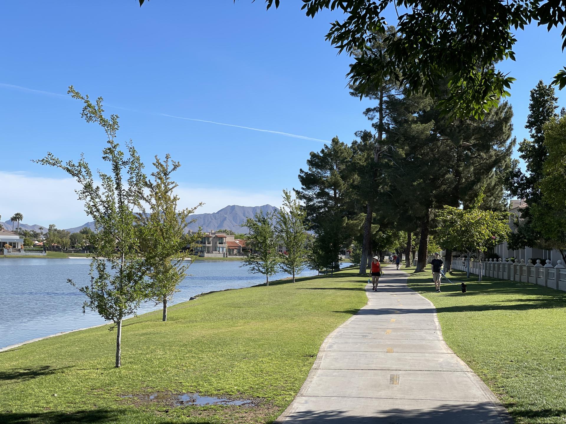 People walk on a paved path beside a lake with trees and mountains.