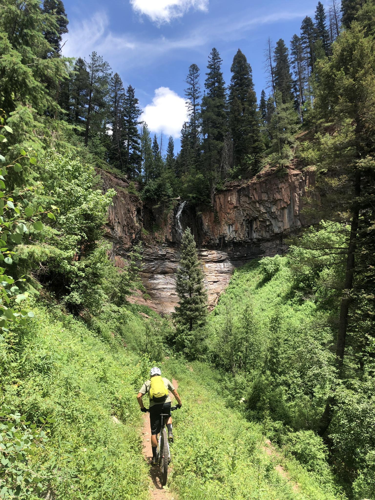 A person bikes on a lush trail towards a waterfall and rocky cliff.
