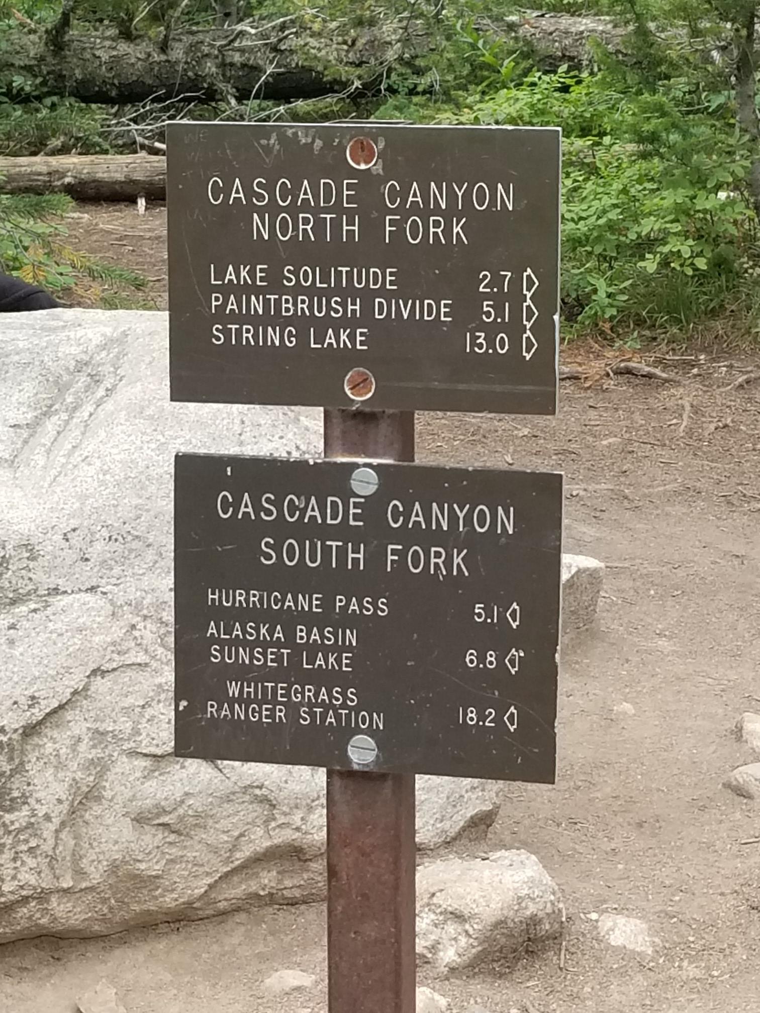 A brown wooden trail sign with white text, indicating various routes and distances.