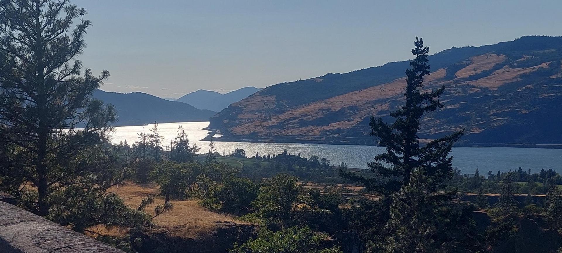Sunny landscape with a wide river reflecting light, surrounded by mountains and trees.
