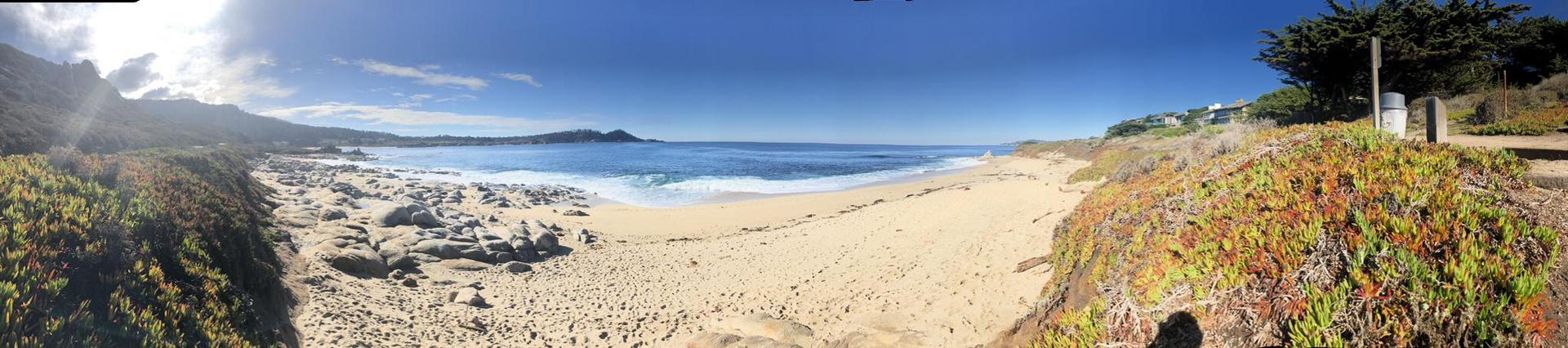 Panoramic view of a sunny sandy beach with waves and rocky coastline.
