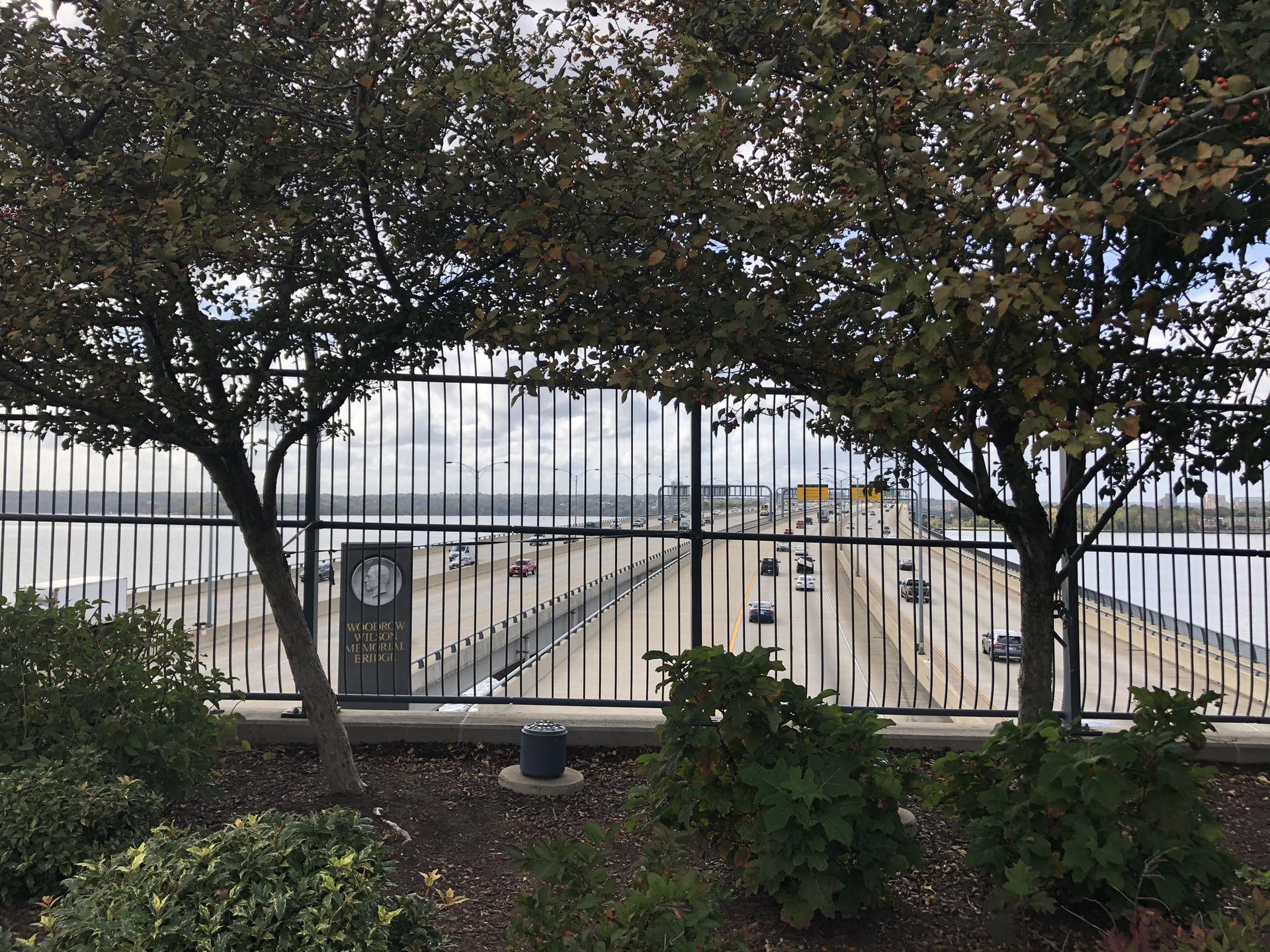 Bridge with cars seen through a black fence and green trees.