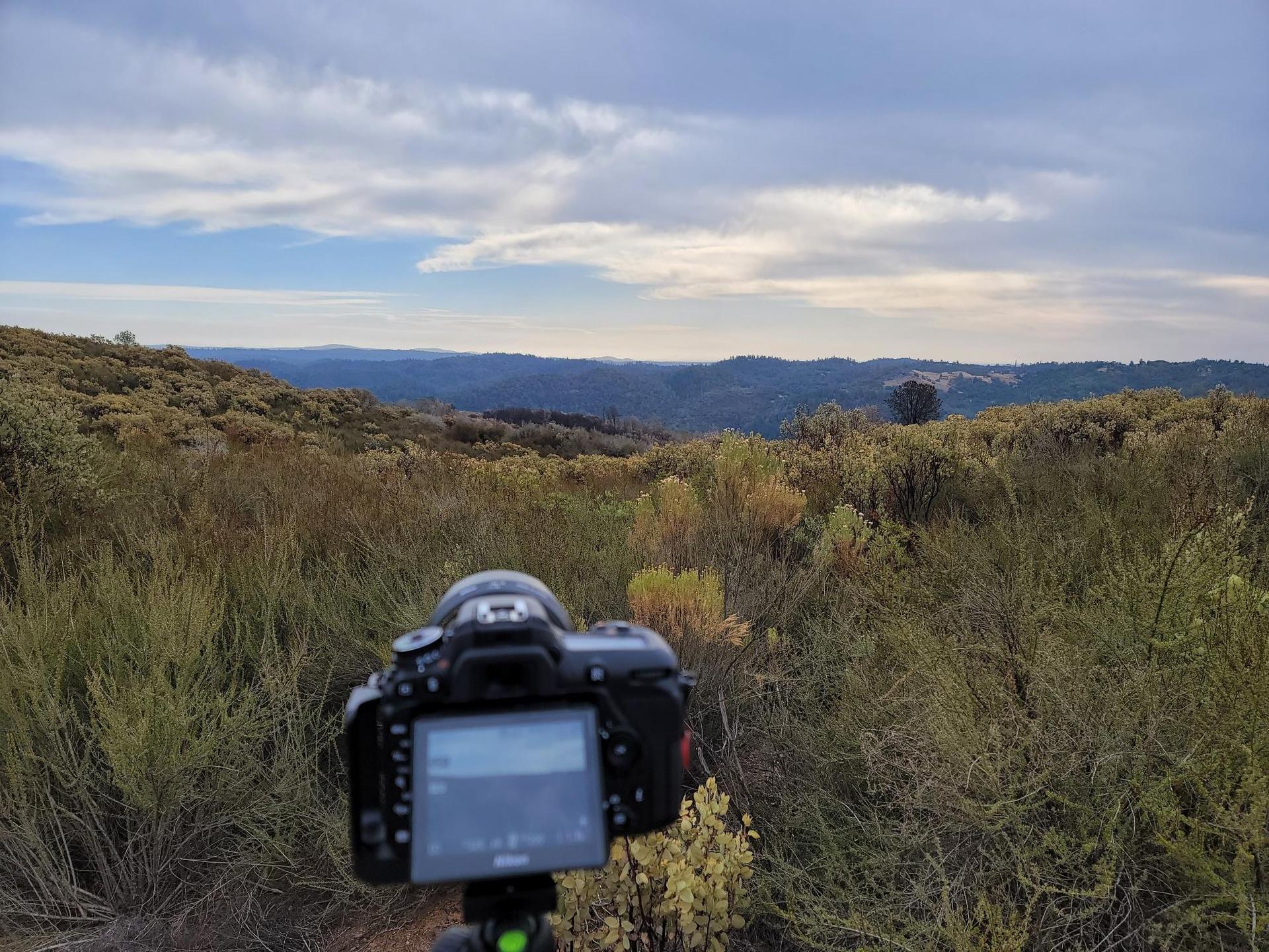 Camera on tripod overlooking a vast, brushy landscape under cloudy skies.