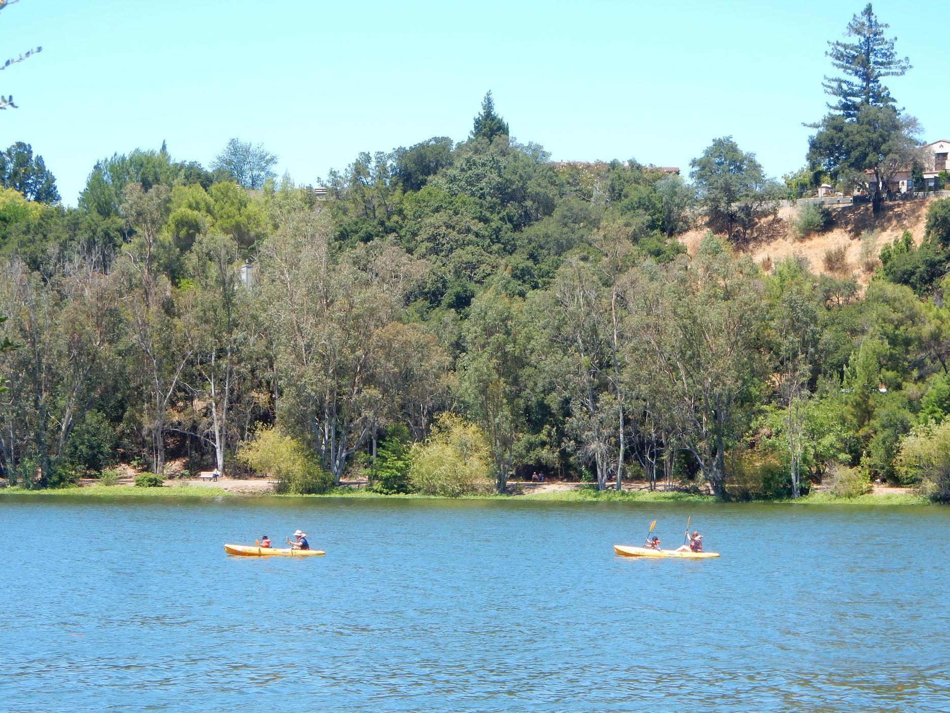 Two people kayaking on a sunny lake with forested hills in the background.