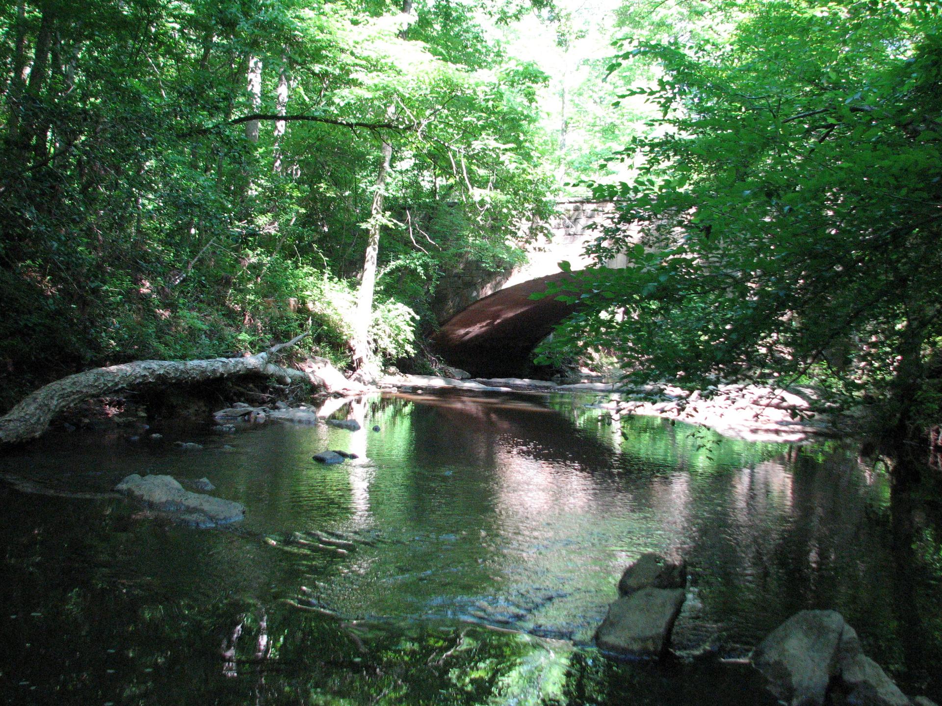 A serene stream flows under a stone bridge, surrounded by green trees.