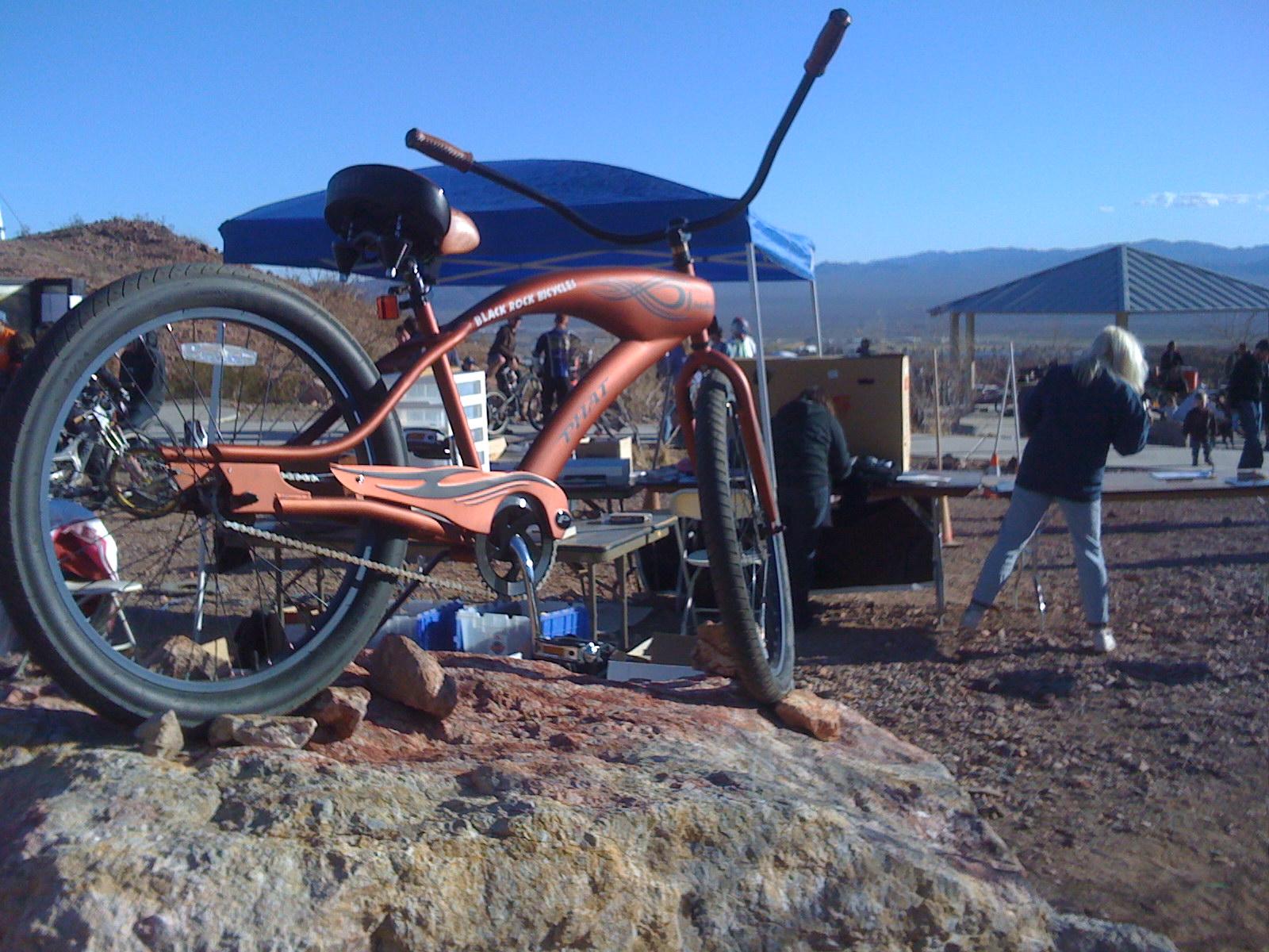Orange cruiser bicycle displayed on rocks with people and tents in background.