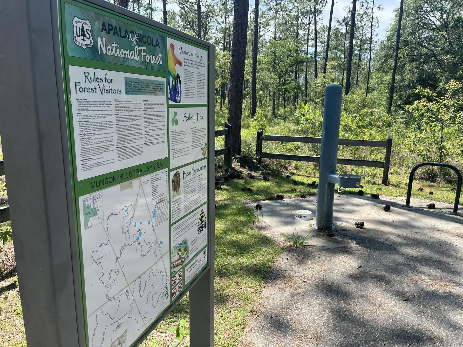 An information sign stands beside a paved area near a forest.