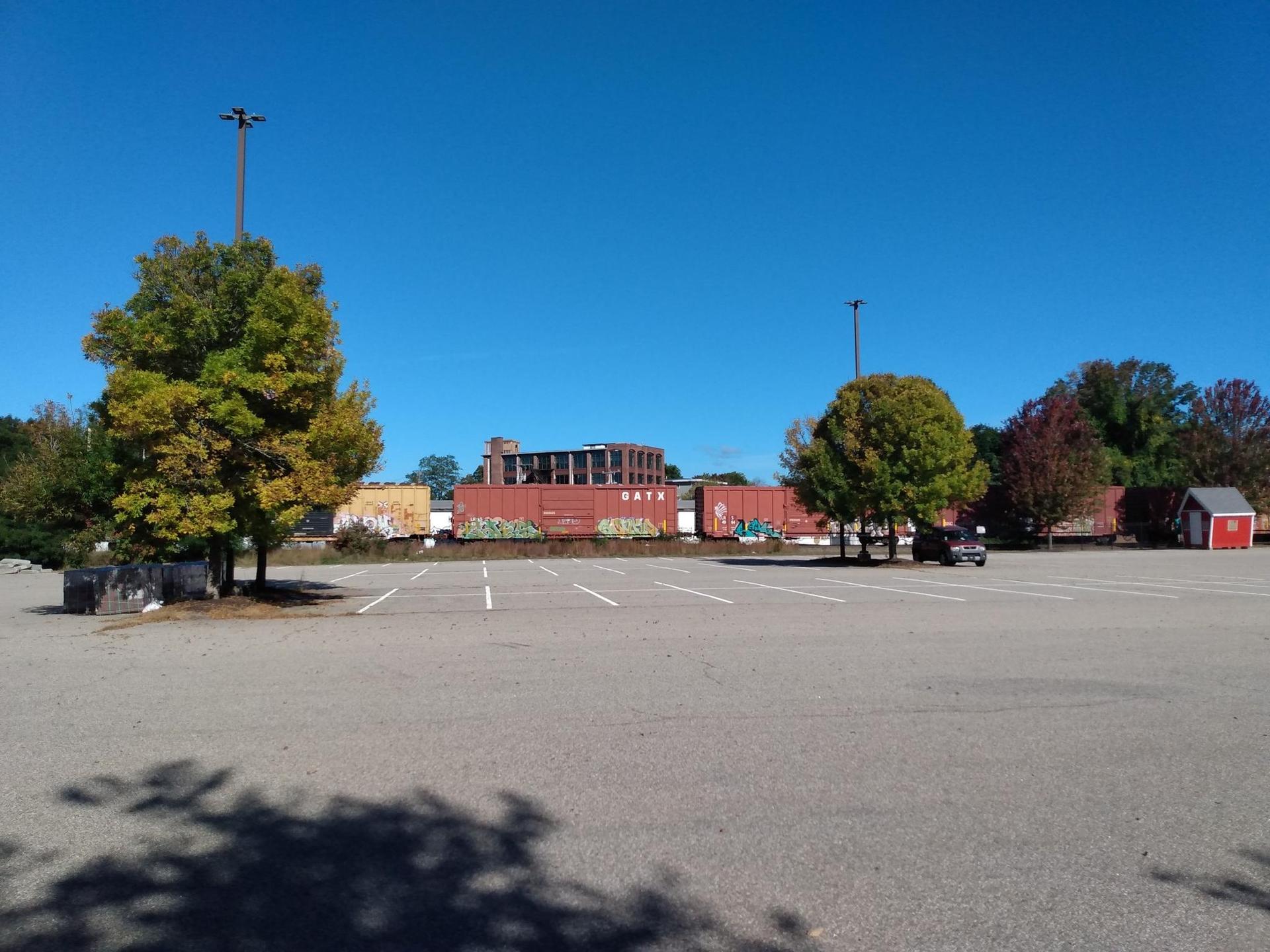Sunny empty parking lot with autumn trees and industrial buildings in the background.