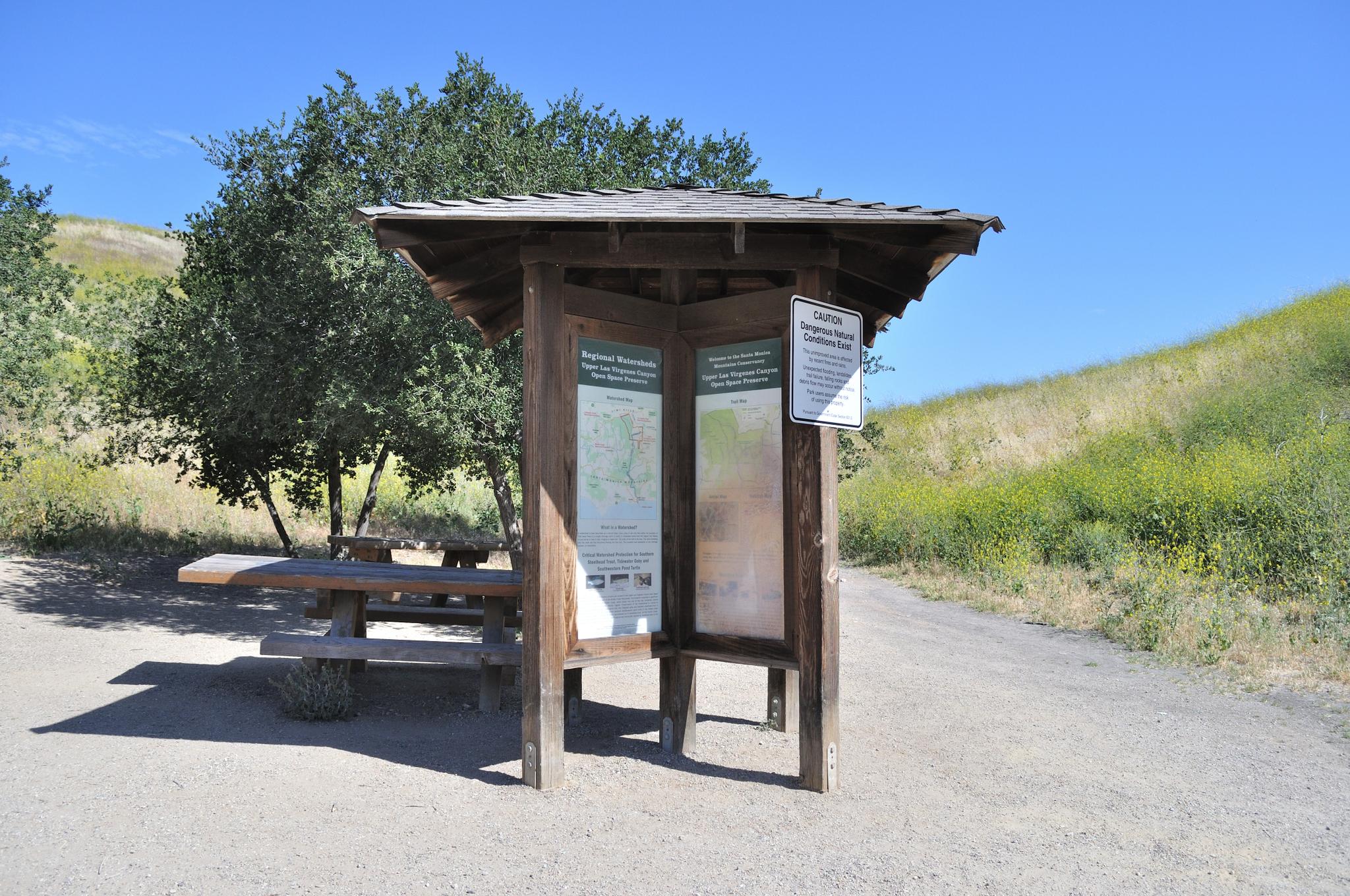 Wooden information kiosk and picnic table next to a dirt path on a sunny day.