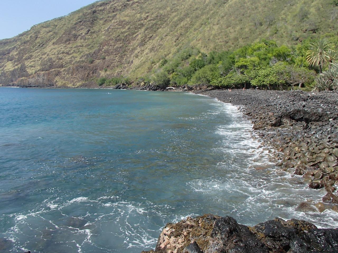 Dark rocky beach meets blue ocean with a steep green hillside under daylight.