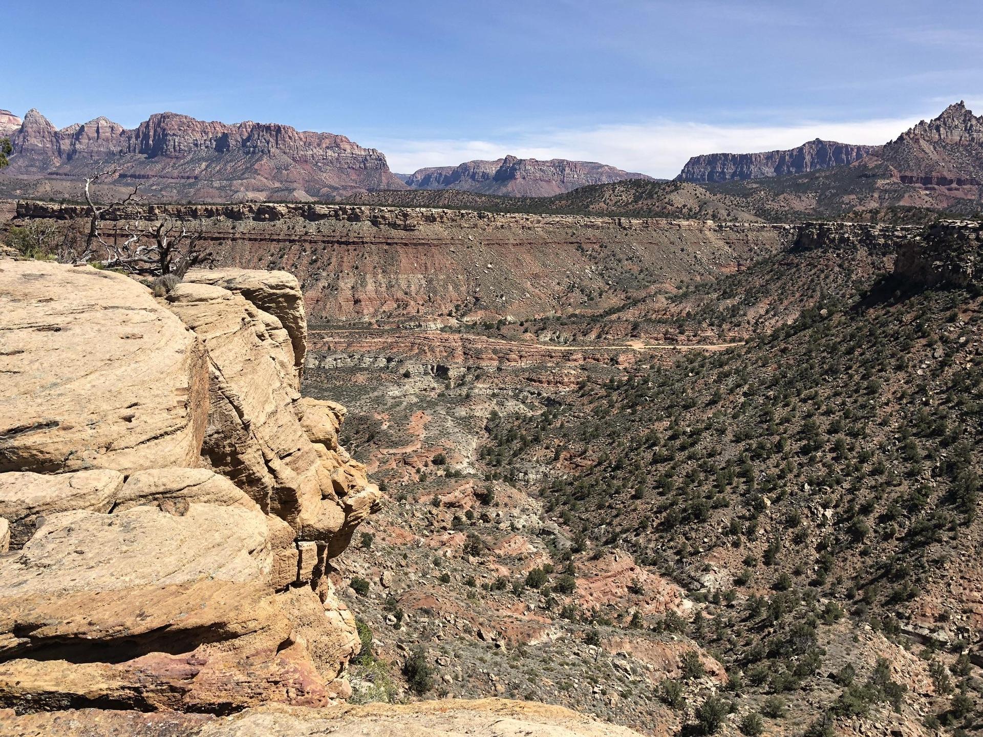 A vast, sunny canyon with layered rock formations and distant mountains under a clear sky.