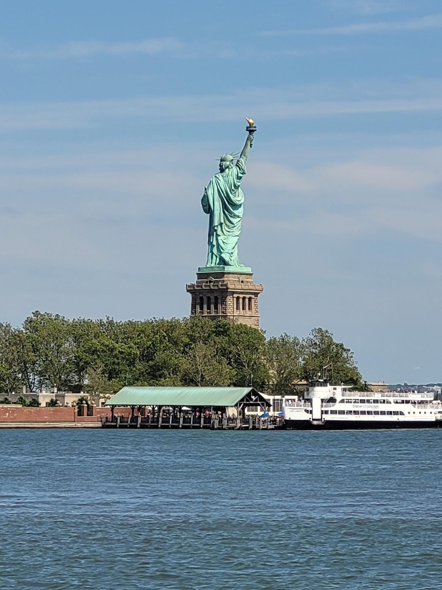 Large green statue on an island with a ferry boat docked nearby.