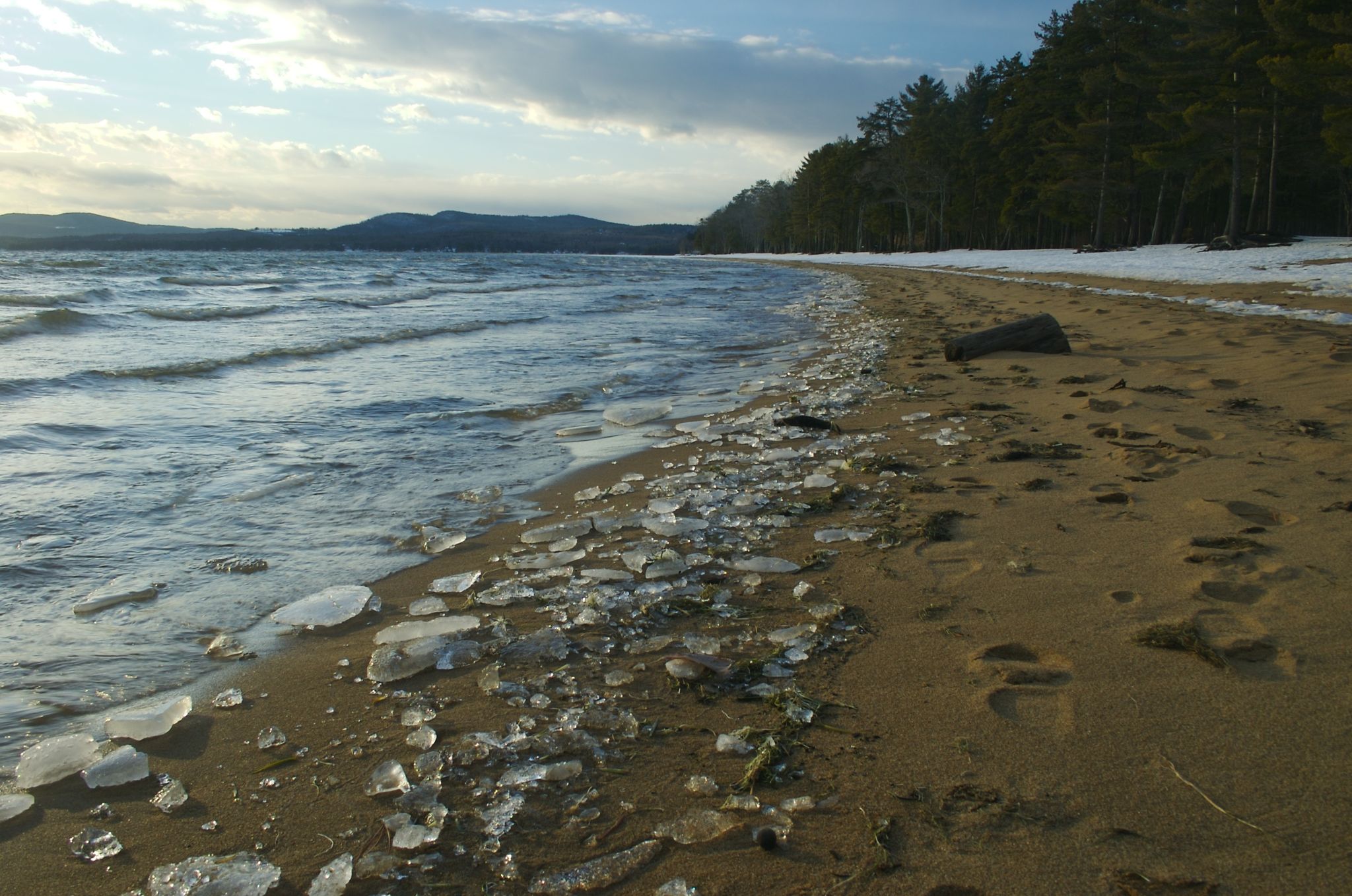 Waves lap a sandy beach with ice and snow under a cloudy sky. - Sebago Lake State Park loop