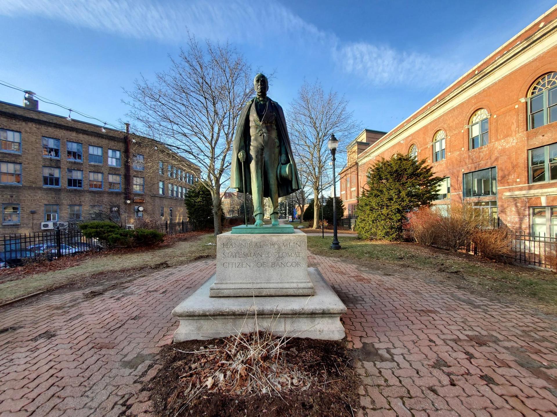 Bronze statue on a stone pedestal in a brick courtyard, sunny.