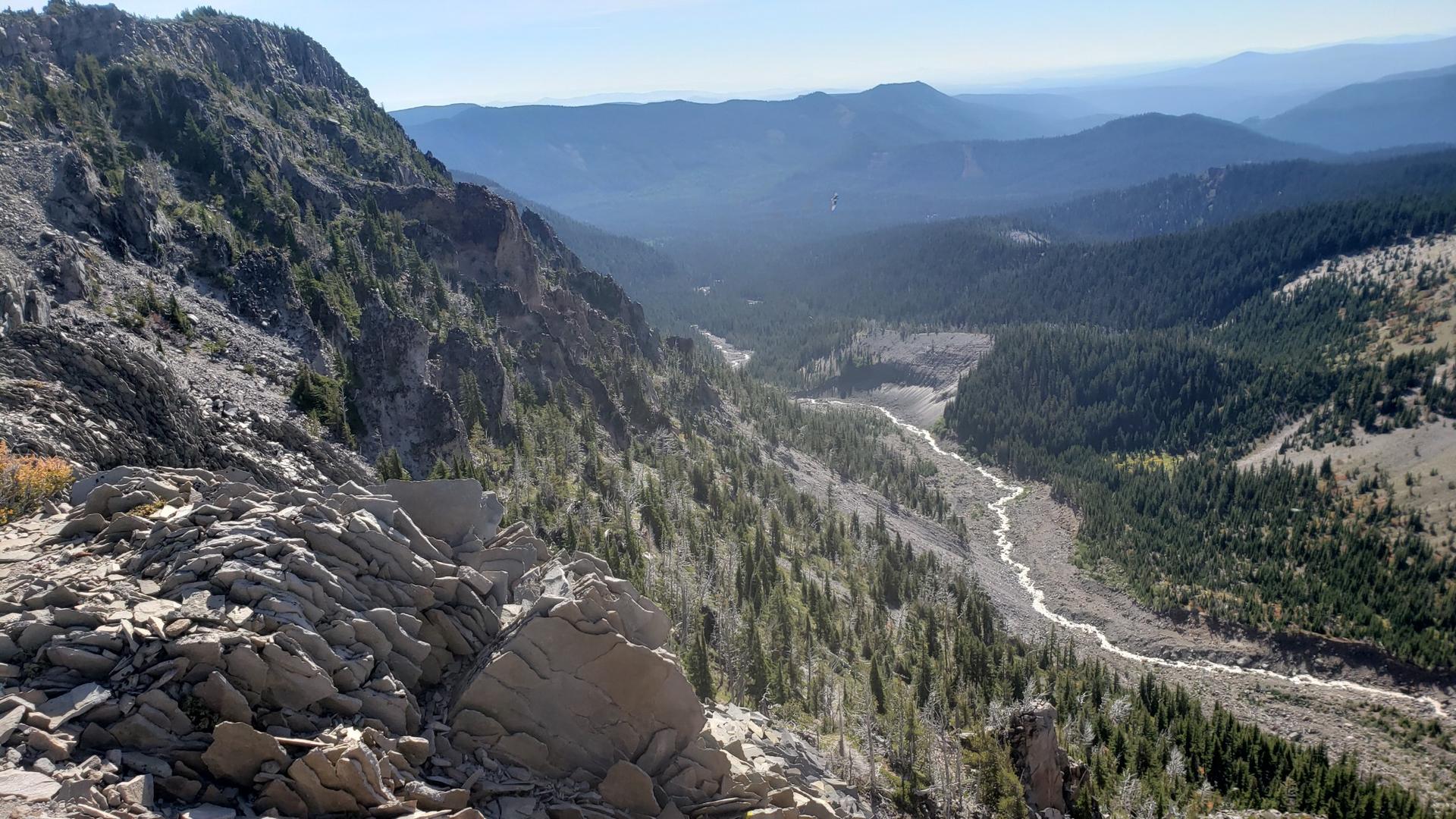 Expansive mountain valley with steep rocky slopes, dense forest, and a wide riverbed.