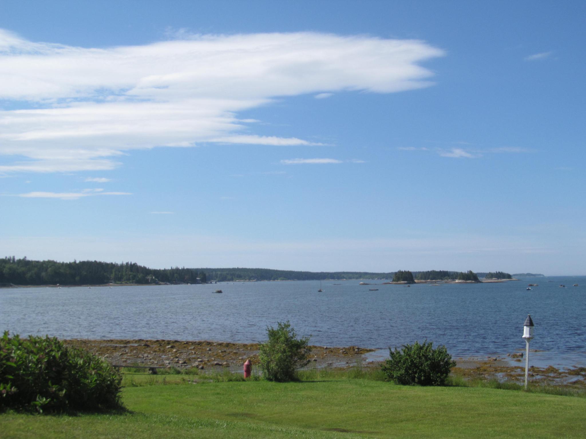 Expansive bay with distant islands, rocky shore, and green grass under blue sky.