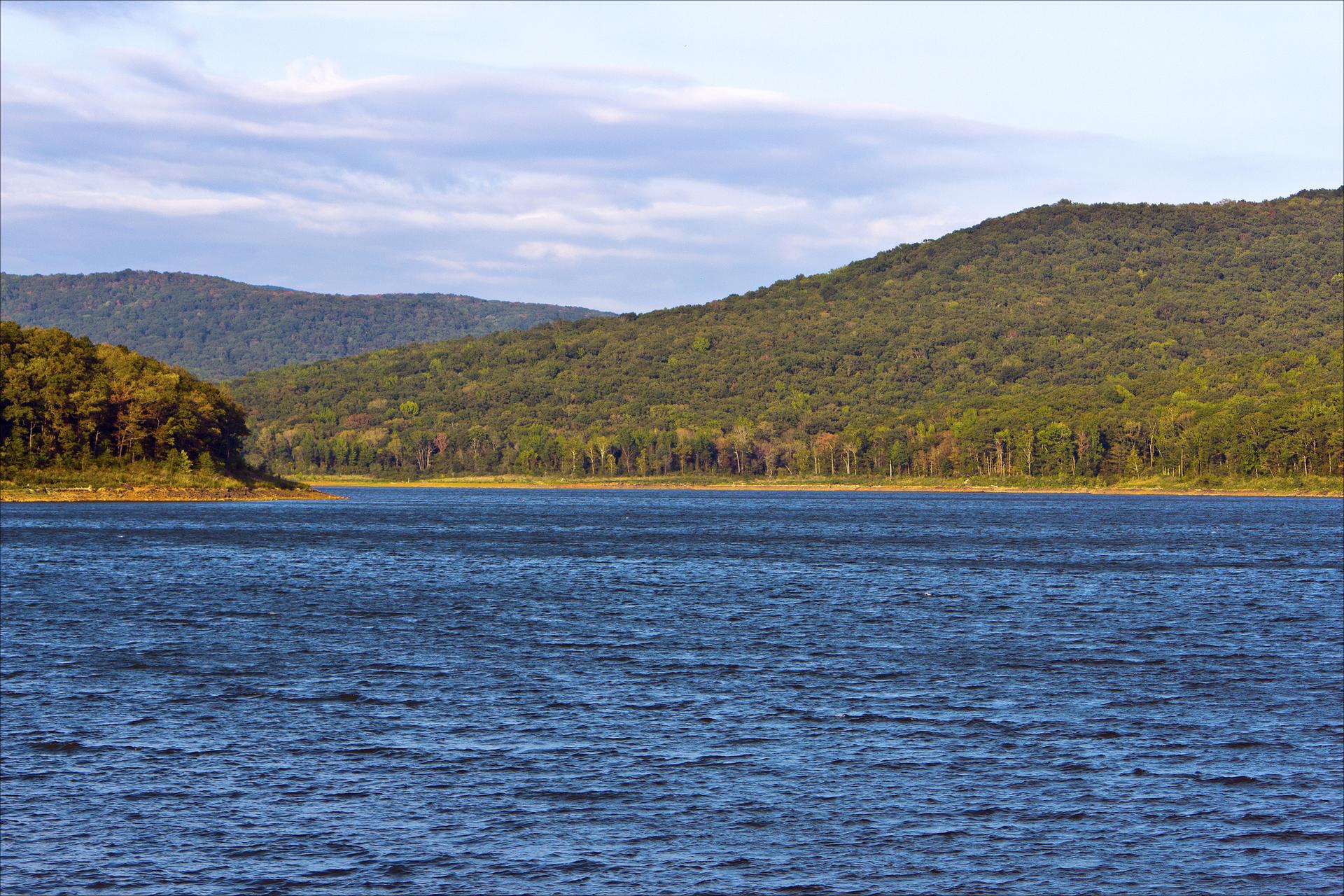 Blue water with ripples, surrounded by green, tree-covered hills under a cloudy sky.