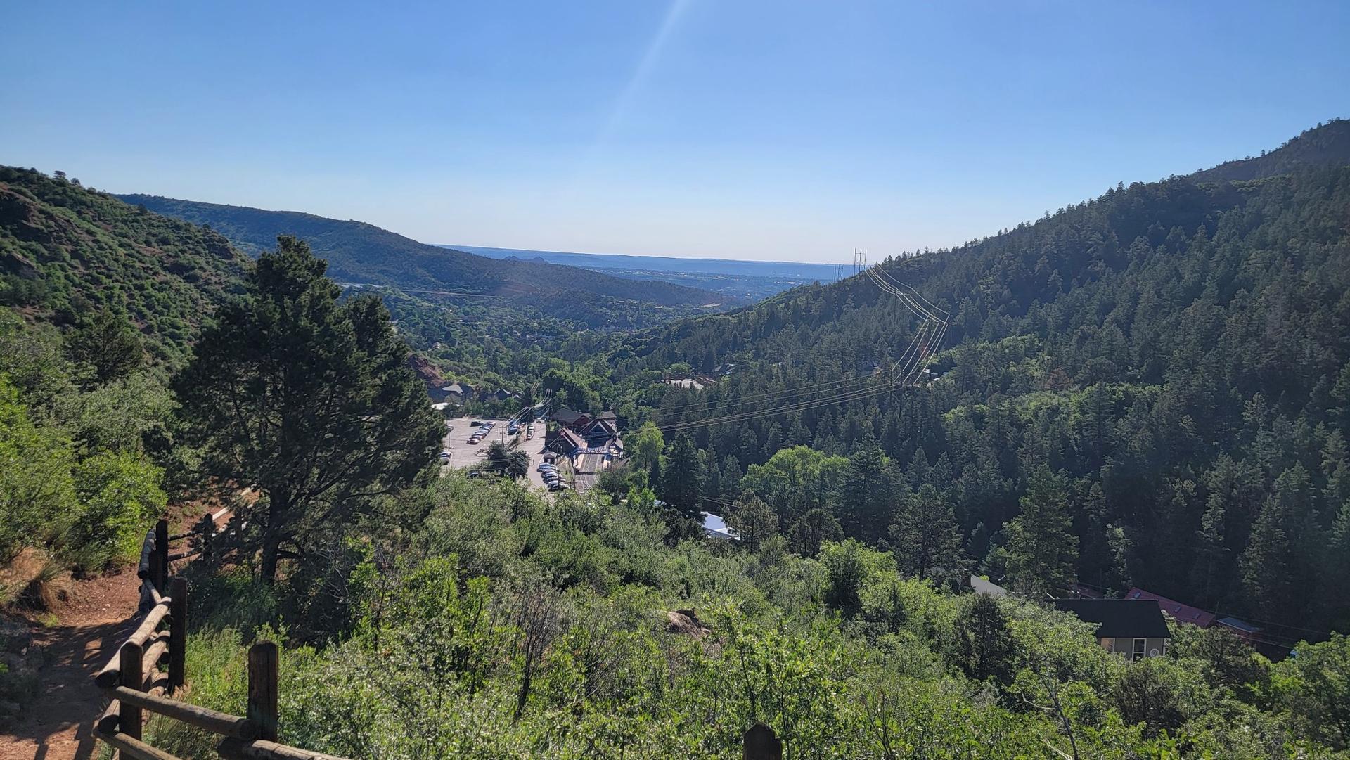 Sunny view of a lush green valley with buildings, surrounded by forested mountains.