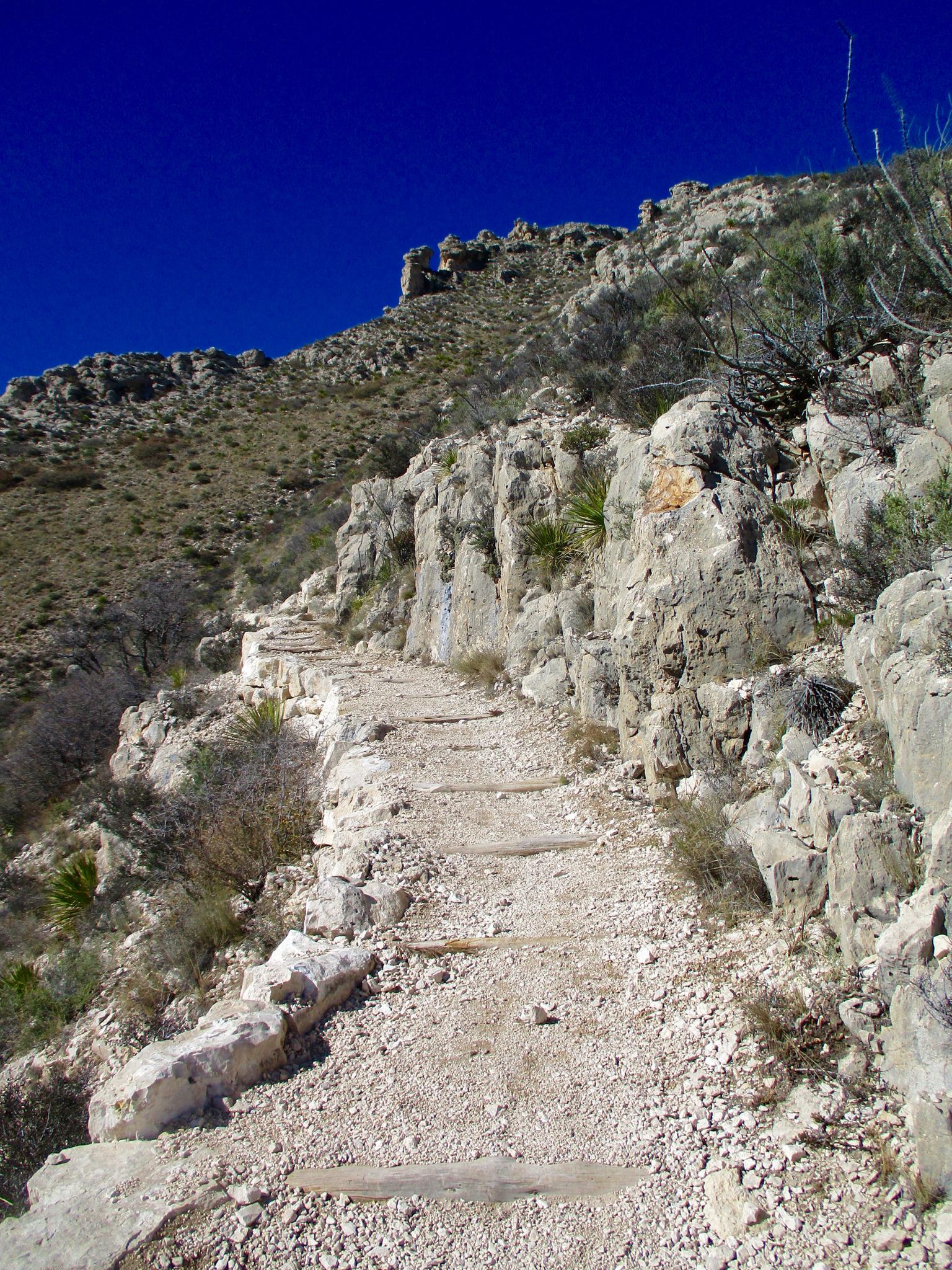 Steep, rocky uphill trail winding through an arid mountain landscape under blue sky.