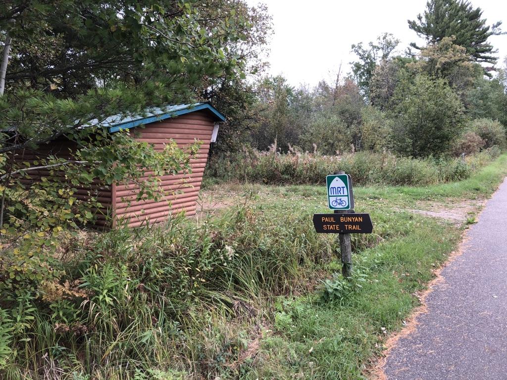 A small wooden shelter and trail sign next to a paved path.