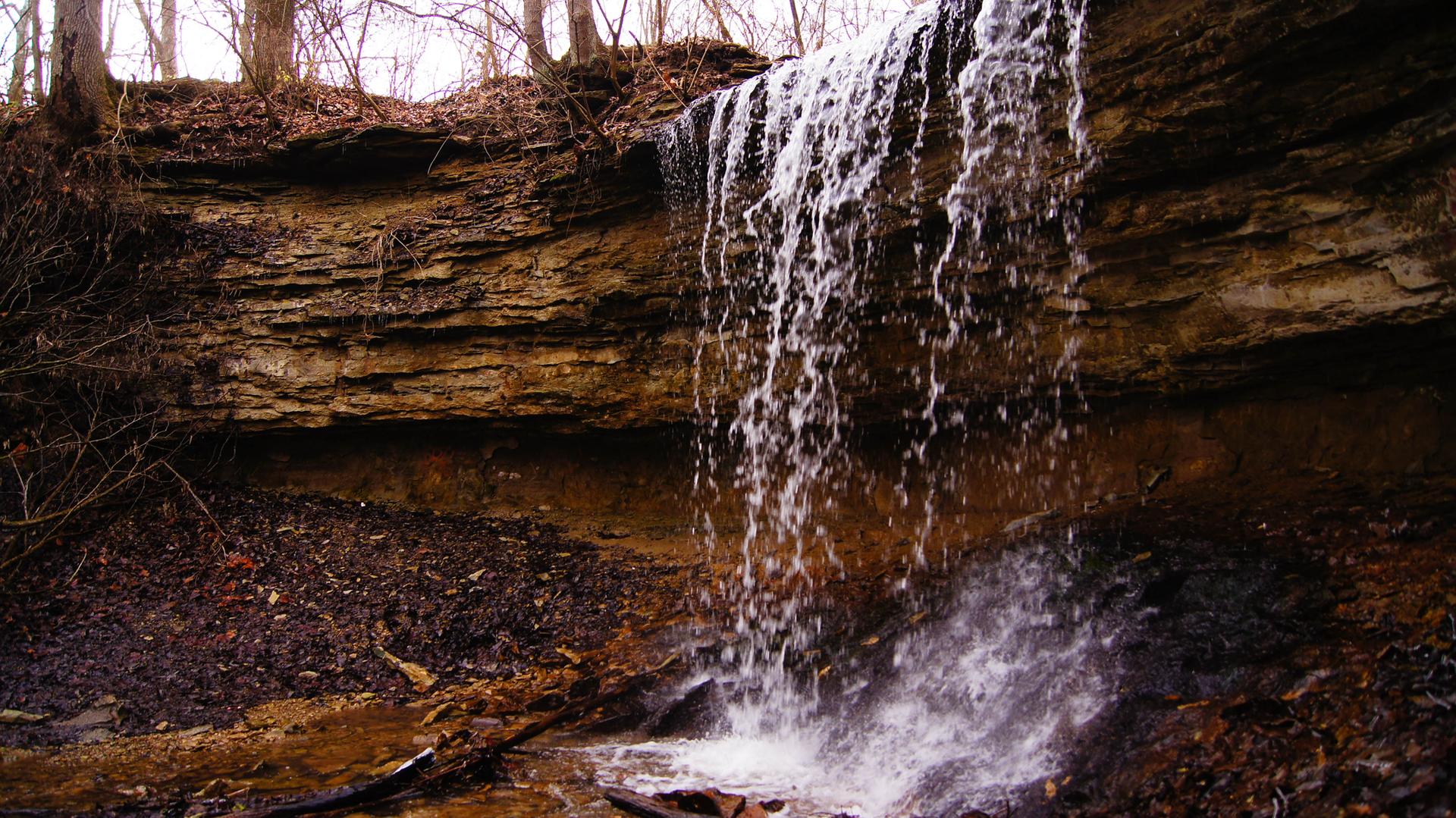 A waterfall cascades over a rocky cliff into a stream, surrounded by bare trees.