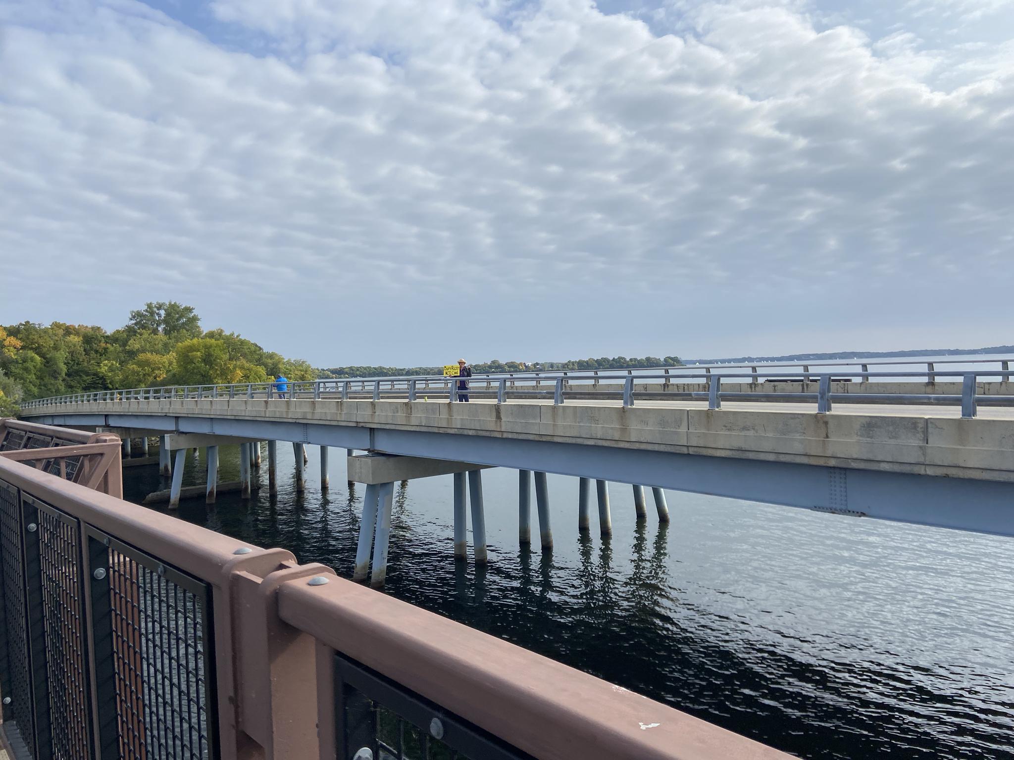 People walk and bike on a long bridge spanning wide water under cloudy skies.