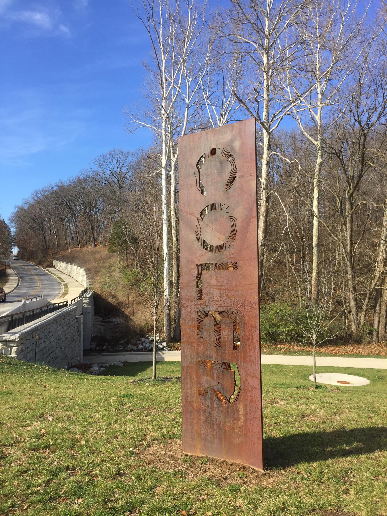 A tall metal sculpture stands in grass with bare trees and a bridge.