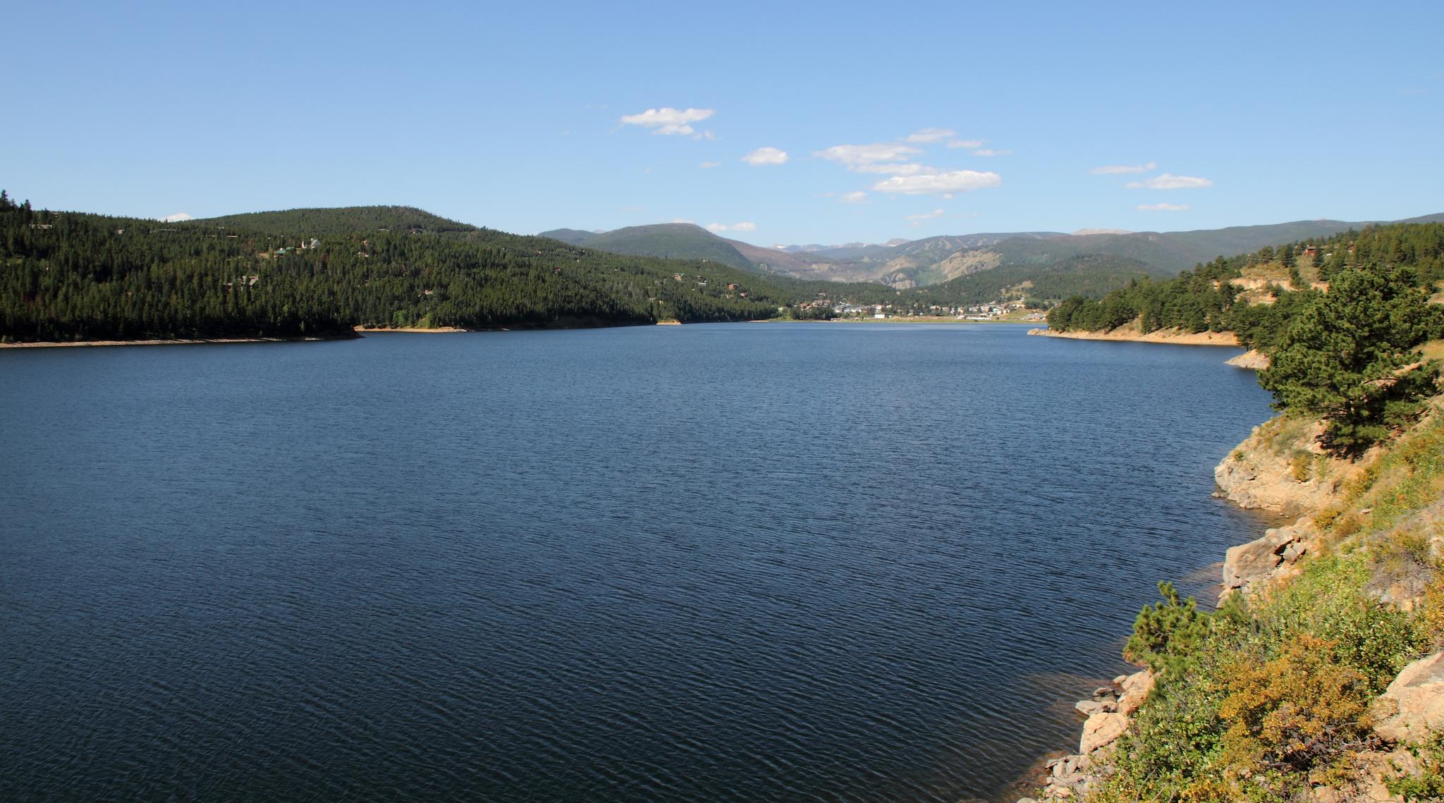 A wide reservoir surrounded by forested hills and a distant town under clear skies.