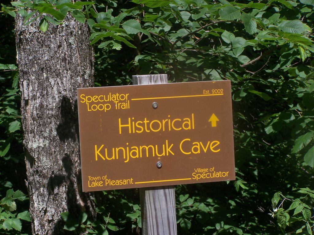 A brown trail sign points to a historical cave, surrounded by green trees in bright sunlight. - Moffitt Beach Campground – Kunjamuk Cave loop from Sacandaga Lake