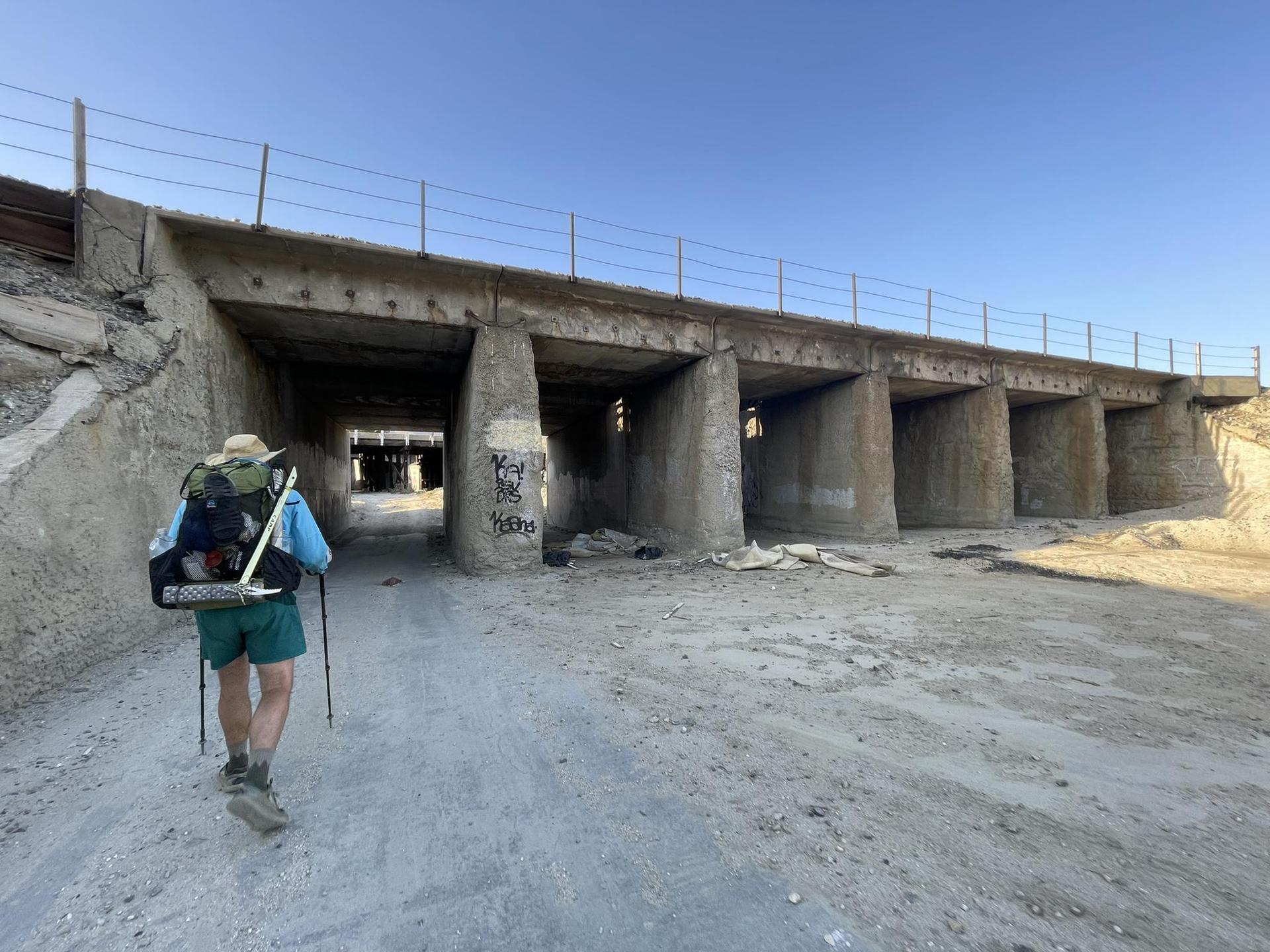 Hiker with backpack and poles approaches a concrete underpass on a dirt path.